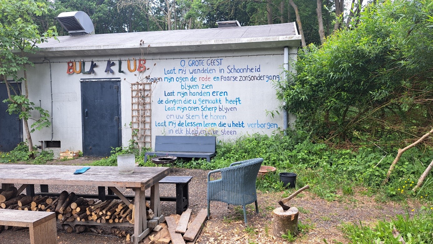Schuur in een bosrijke omgeving met een kleurrijk bord "BUK KLUB" boven de deur en een blauwe tekst op de muur. Voor de schuur staan een houten tafel met houtstapels eronder, een stoel en een hakblok met een bijl. Omringd door bomen en struiken.