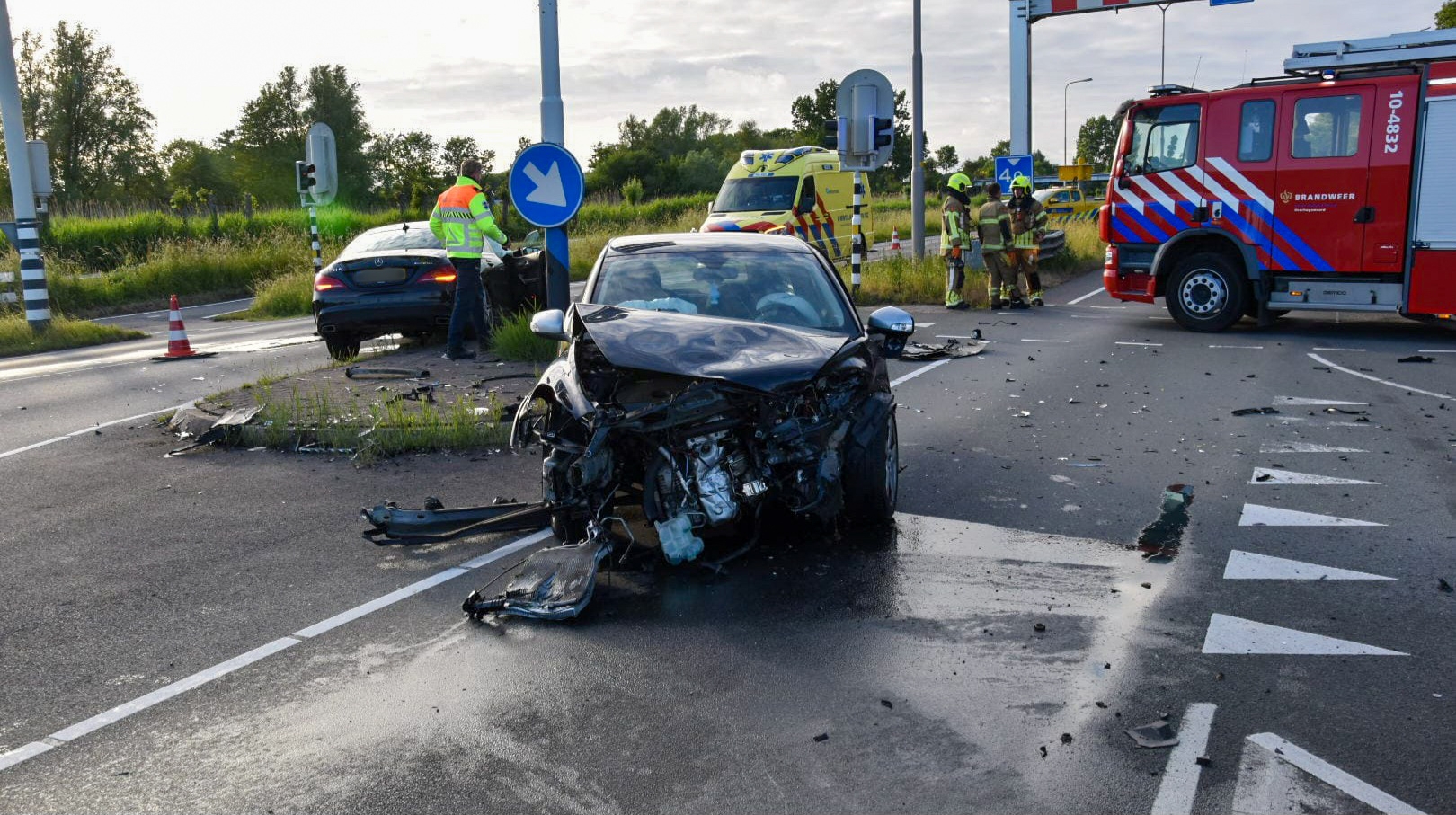 Twee auto's met aanzienlijke schade na een botsing op een kruispunt, met een politiewagen, brandweerwagen en ambulance ter plaatse, omgeven door puin op de weg.