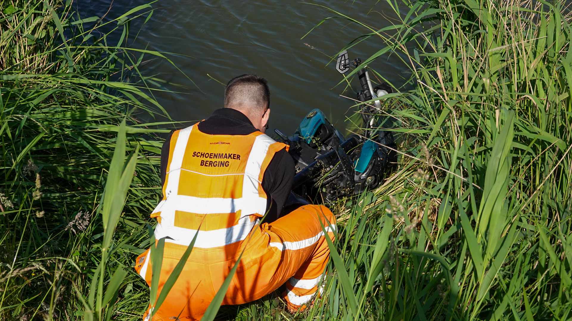 Persoon in oranje veiligheidskleding van "Schoenmaker Berging" buigt zich voorover naar een scooter in het water, omringd door hoge grassen.