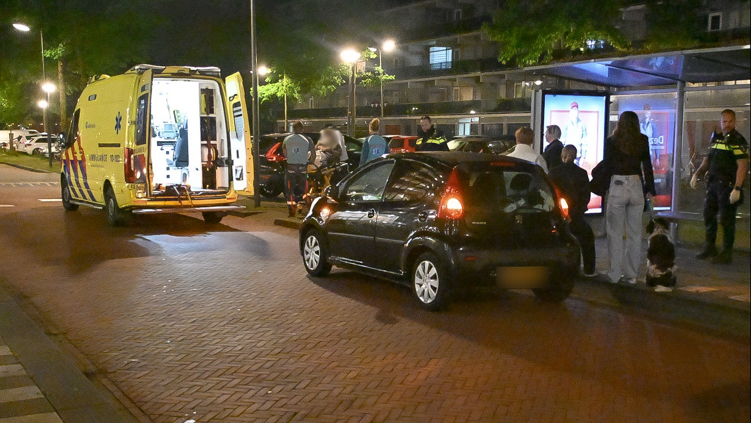Nachtelijke scène met een gele ambulance met open achterdeuren, geparkeerd nabij een zwarte auto op een straat. Er zijn mensen, waaronder politieagenten, bij een bushalte in gesprek. De omgeving is verlicht door straatlantaarns.