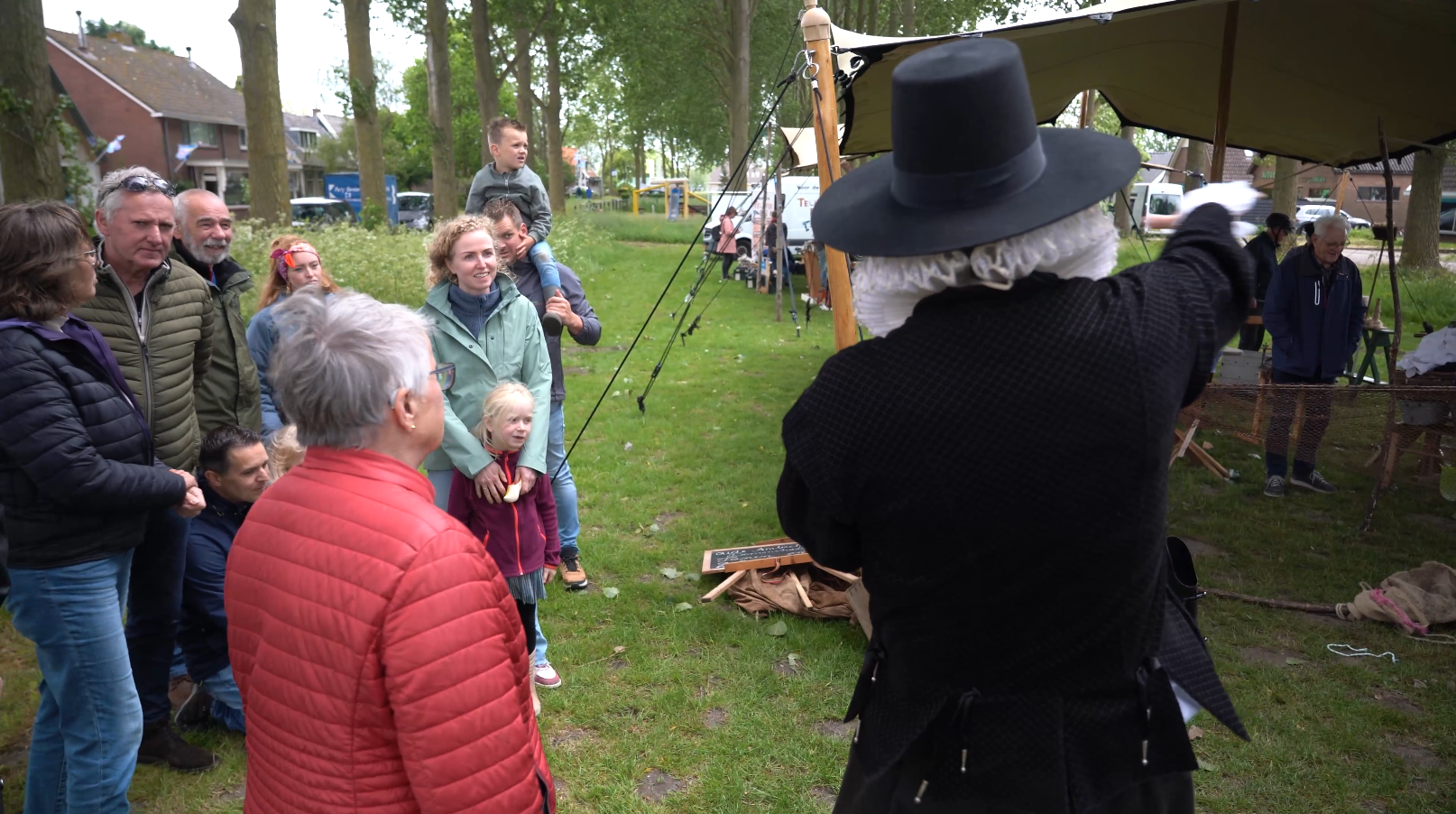 Groep mensen kijkt naar een persoon met een zwart kostuum en hoed op een buitenlocatie onder een tentachtige structuur.