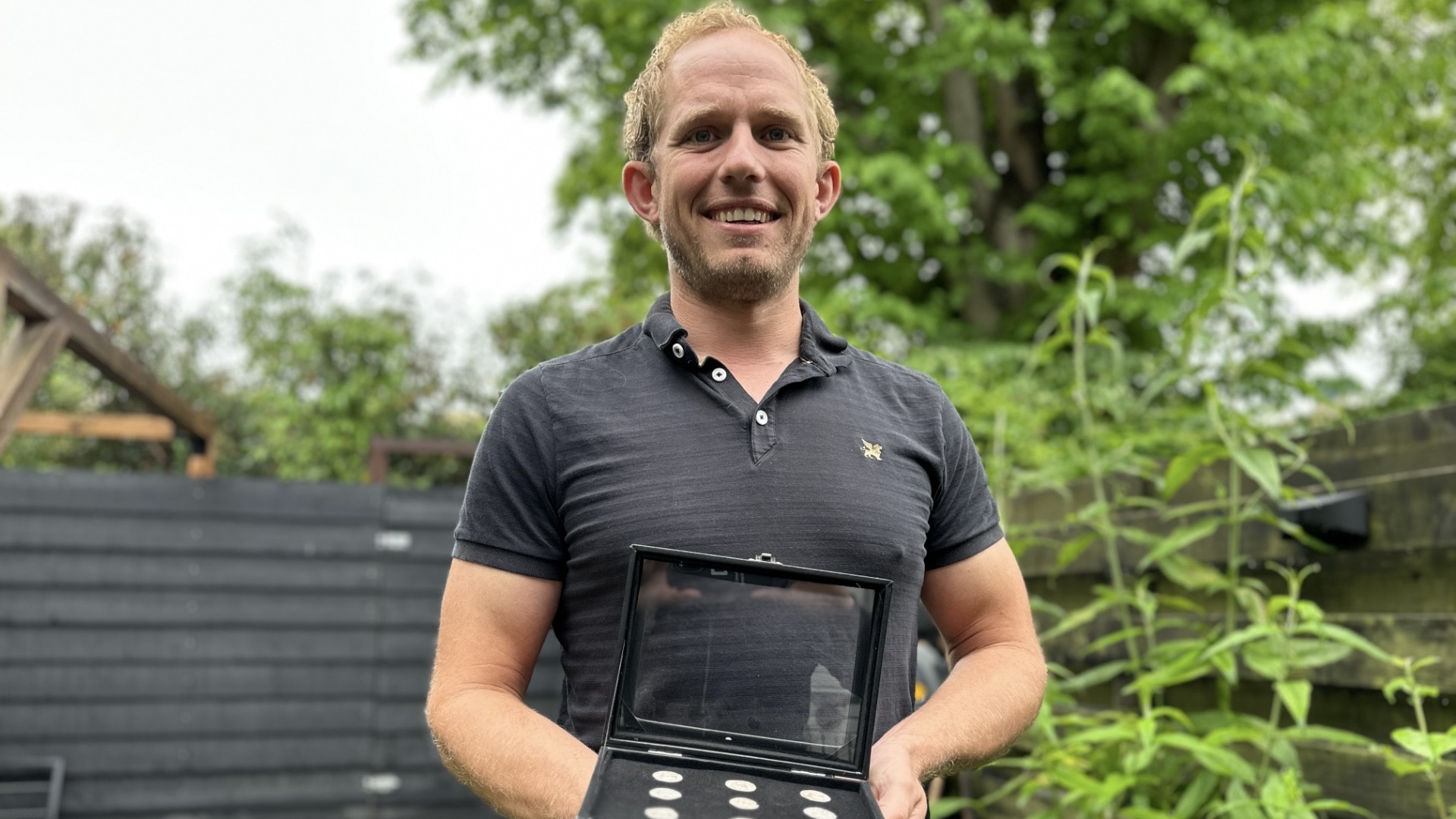 Man in a black shirt holding a display case with coins, standing in a garden with greenery in the background.