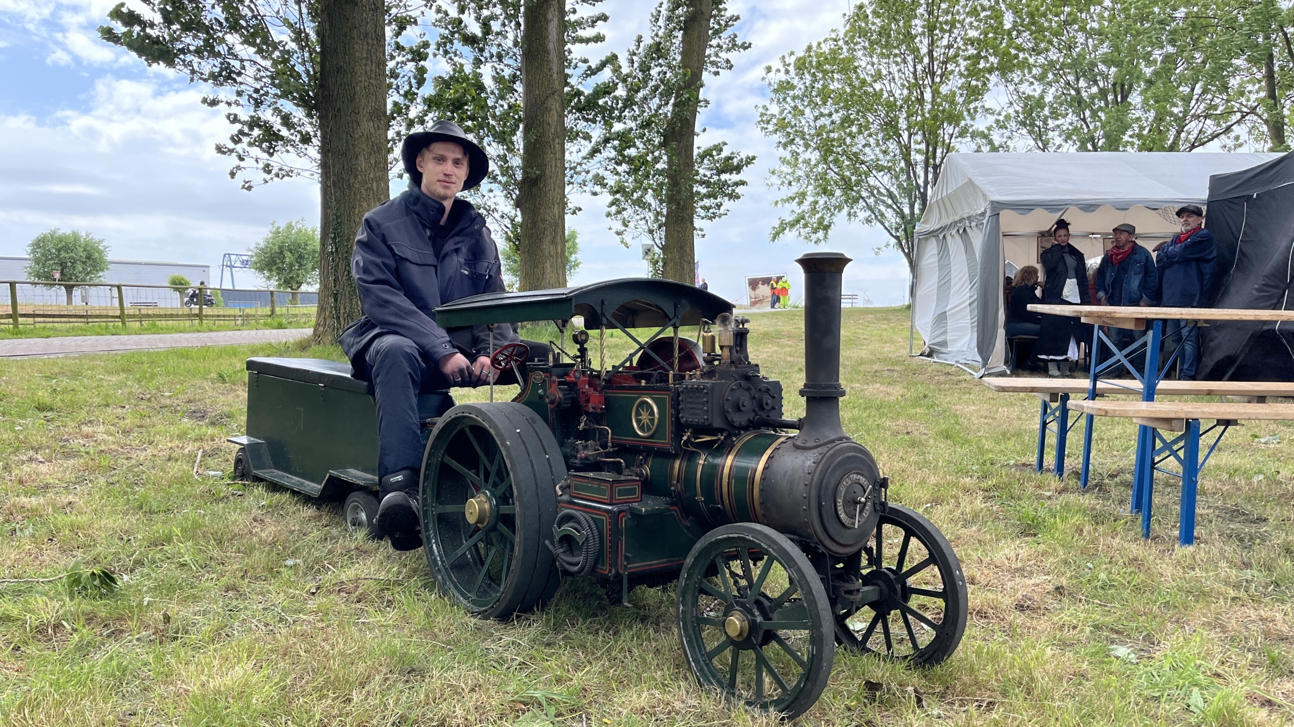 Een man zittend op een miniatuur stoomtractor naast een tent in een grasveld, omringd door bomen.