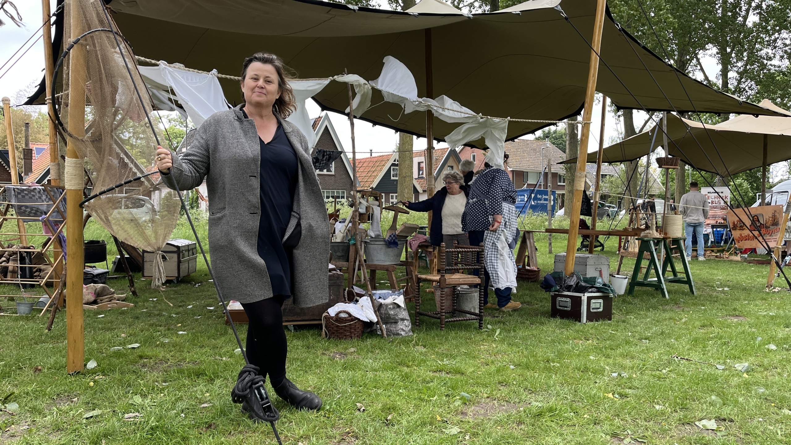 Een vrouw in een grijze jas loopt op een grasveld met op de achtergrond een marktkraam met doeken en mensen die bezig zijn met verschillende activiteiten in een buitenomgeving.