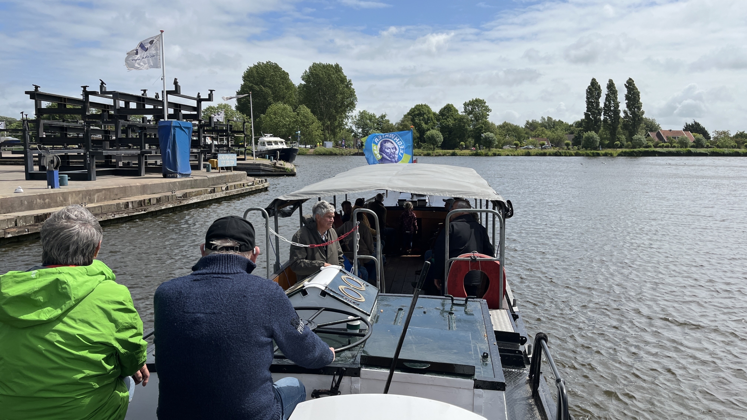 Een groep mensen in een boot varen over een rivier, met een landschap van bomen en huizen op de achtergrond en een vlag met een afbeelding op het dak van de boot.