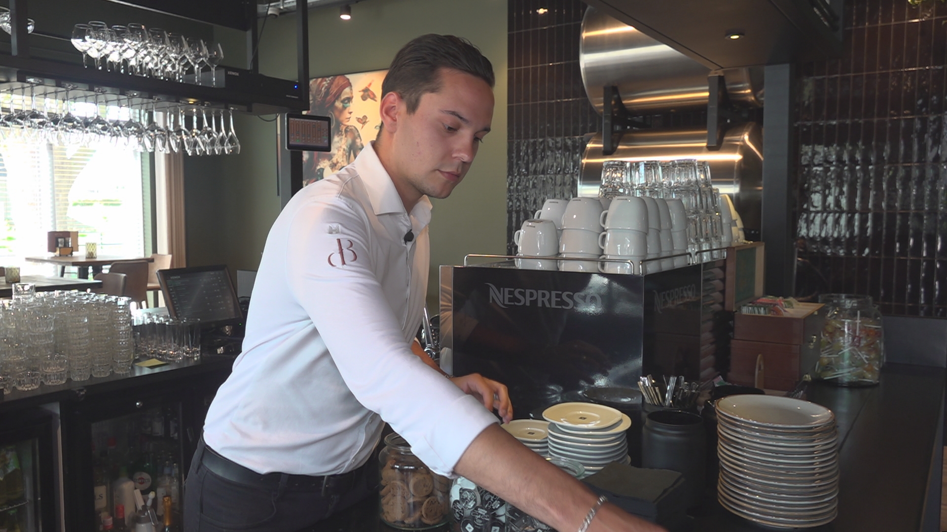 Een barman in een wit overhemd werkt achter de bar met glazen en kopjes opgestapeld naast een Nespresso-machine.