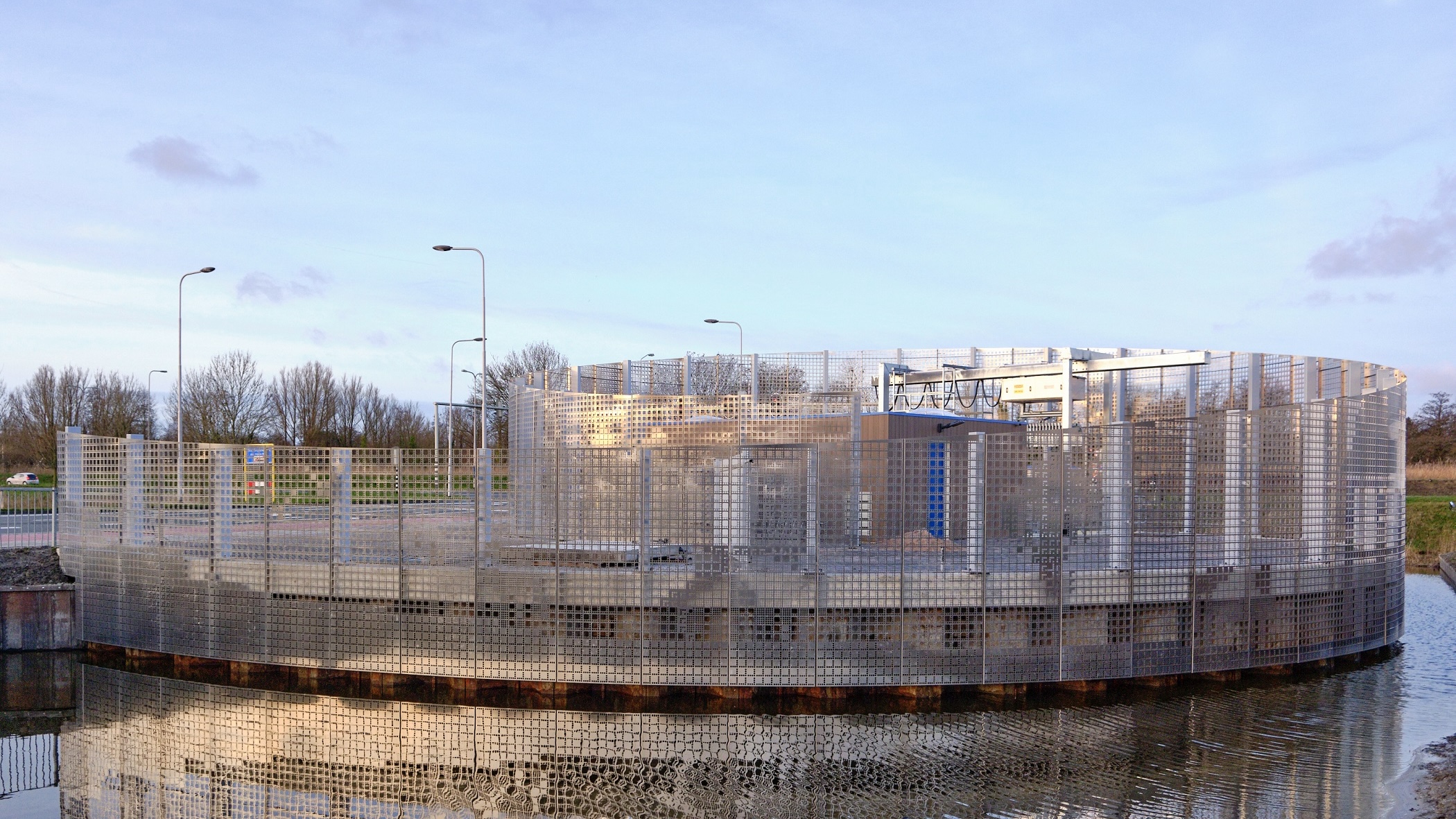 Een ronde structuur met een rastervormig metalen hekwerk, gelegen aan de rand van een waterpartij, onder een heldere hemel.