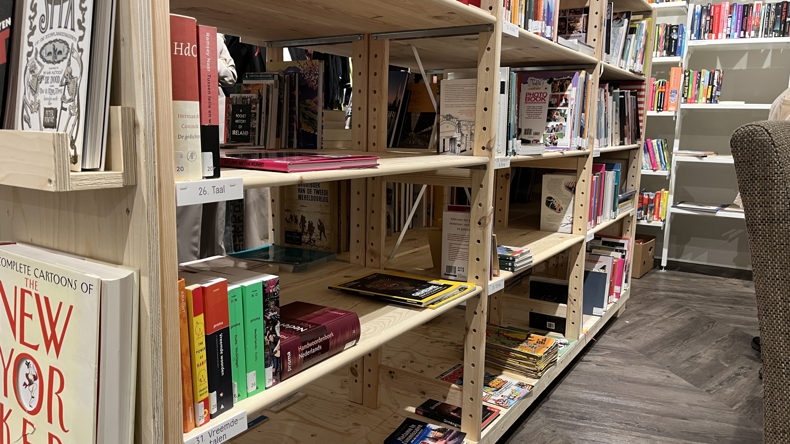 Planken met boeken in een houten boekenkast in een boekwinkel, met daarin diverse boeken in verschillende formaten en kleuren. Op de achtergrond zijn meer boekenkasten zichtbaar, evenals een fauteuil aan de rechterkant.