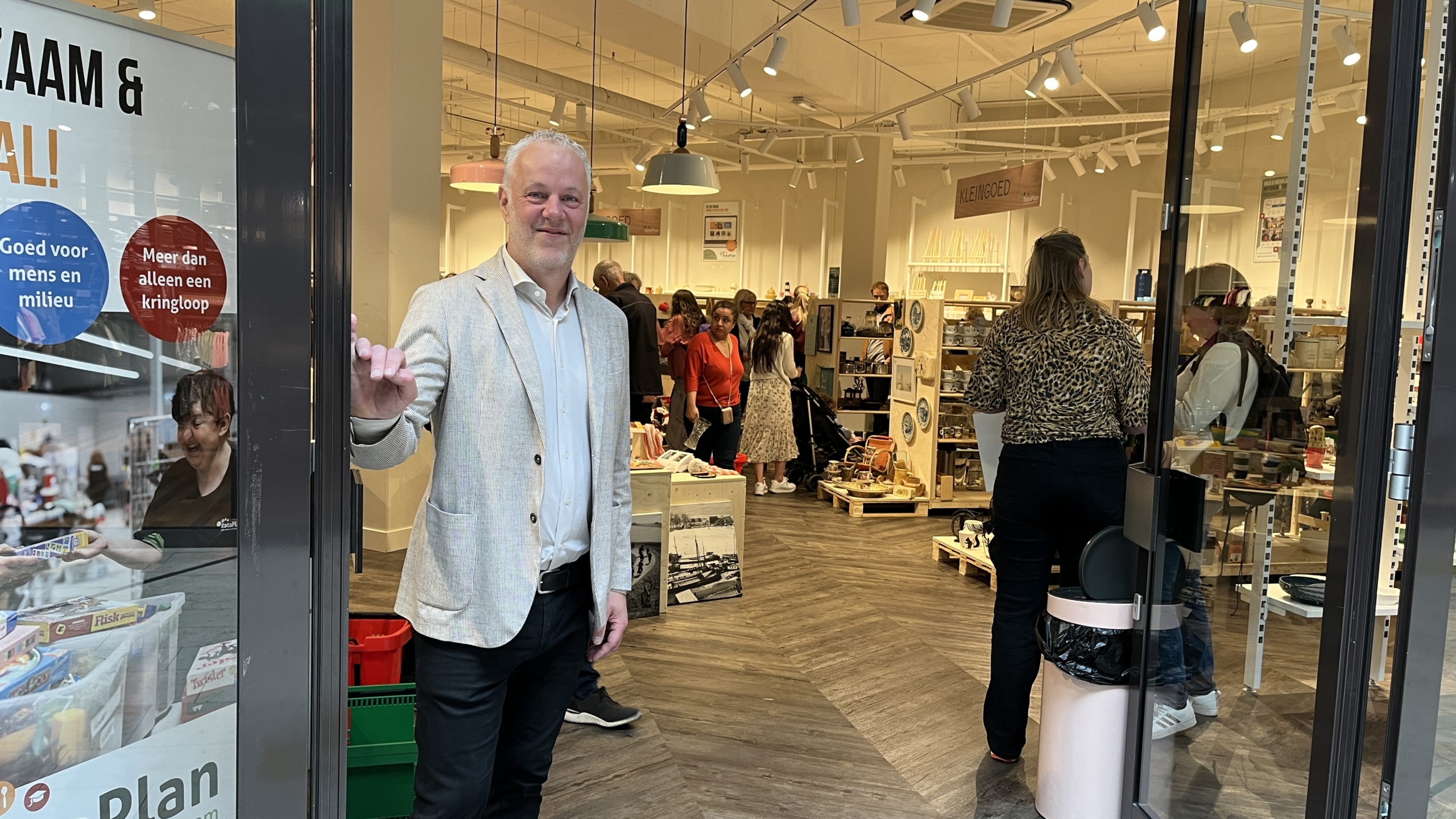 Een man in een lichtgekleurde blazer staat in de deuropening van een winkel met mensen binnenin. De winkel heeft houten vloer en is gevuld met diverse artikelen. Een groot bord naast de deuropening heeft teksten in het Nederlands over duurzaamheid en kringloop.