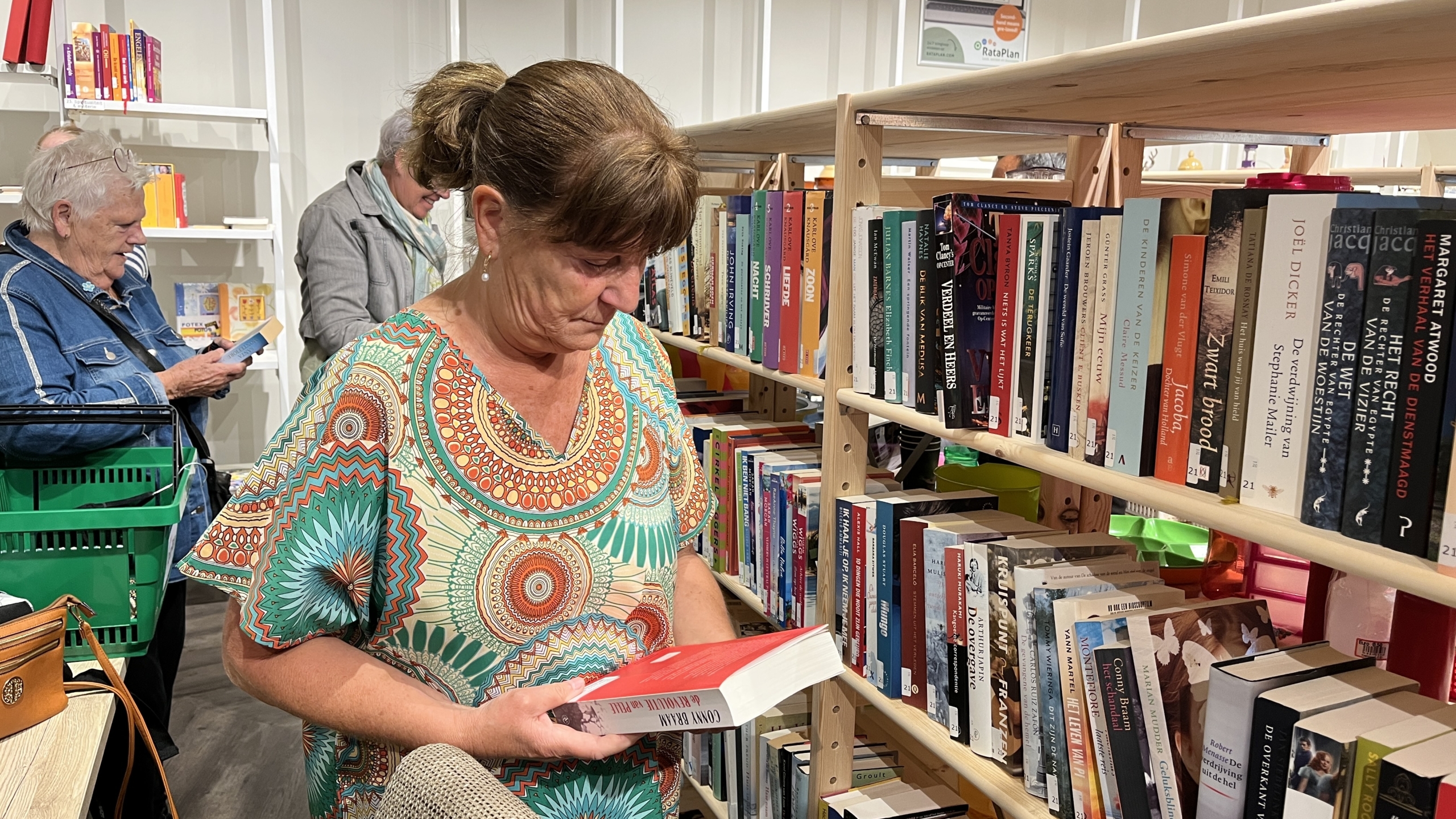 Een vrouw in kleurrijke kleding bekijkt een boek voor een boekenplank in een winkel, terwijl andere mensen in de achtergrond ook bezig zijn met boeken in een winkelomgeving.