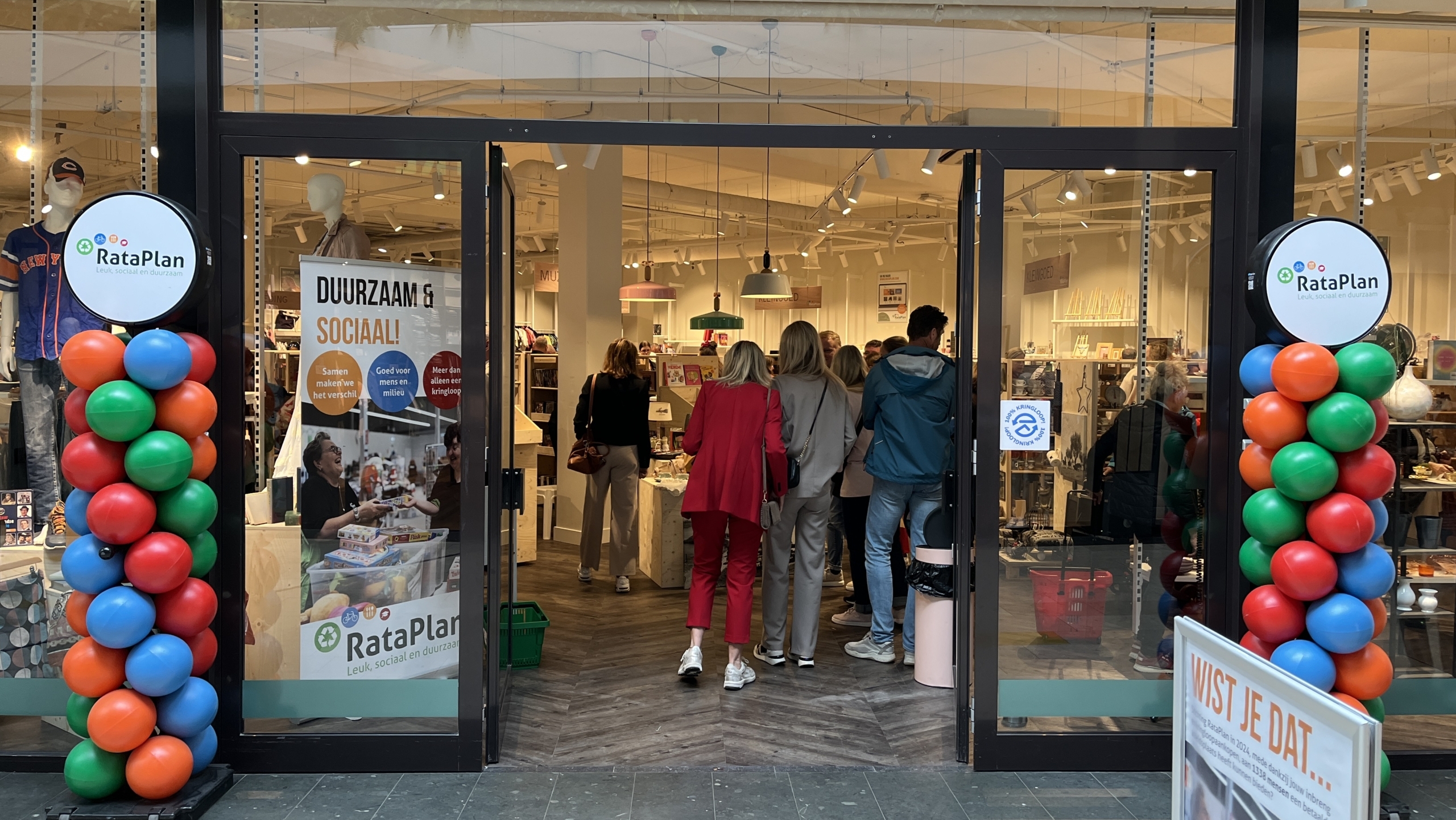 Ingang van een kringloopwinkel genaamd RataPlan, versierd met kleurrijke ballonnen, waarbinnen mensen rondlopen en winkelend bezig zijn.