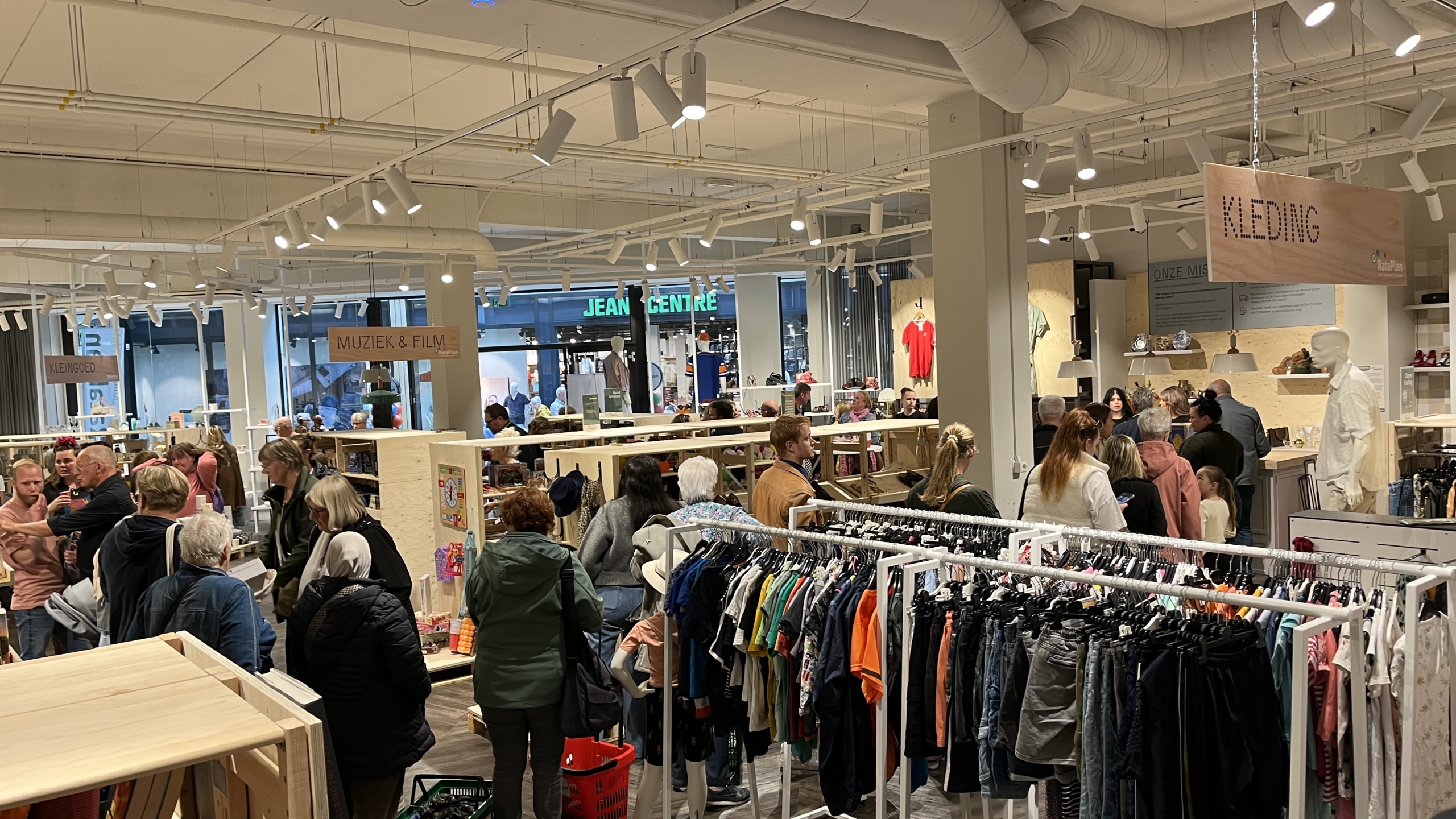 Een drukbezochte winkel interieur met verschillende afdelingen zoals kleding, muziek en film, en kleingoed, met veel mensen die door de gangpaden lopen en aan het winkelen zijn.