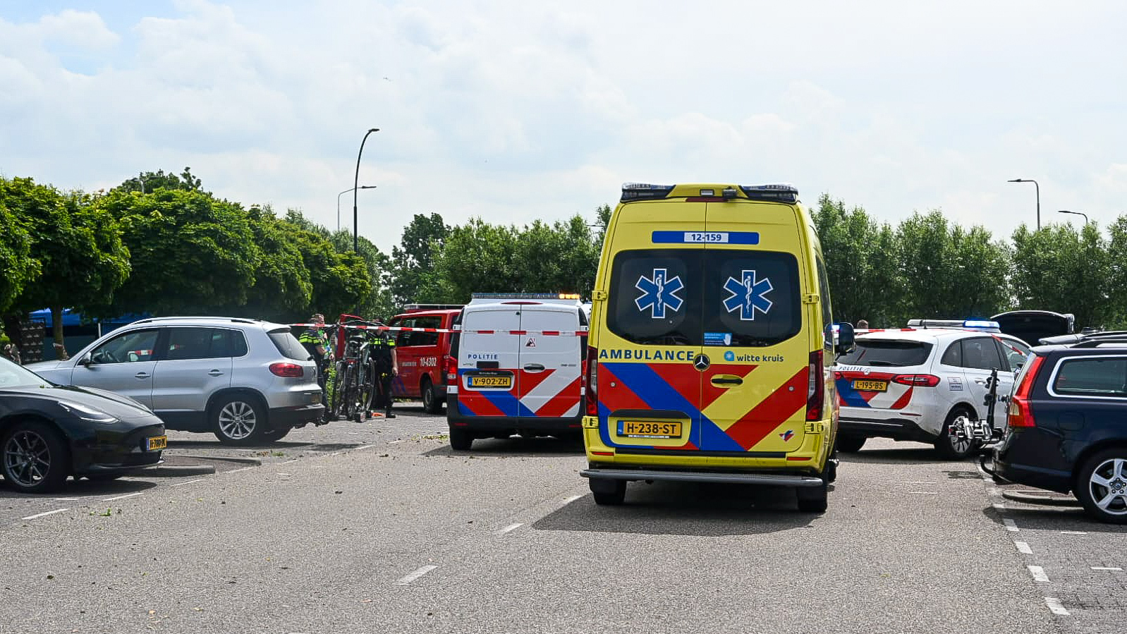 Reddingsvoertuigen, waaronder een ambulance en twee politieauto's, staan opgesteld op een parkeerplaats, omringd door andere auto's en groen, op een bewolkte dag.