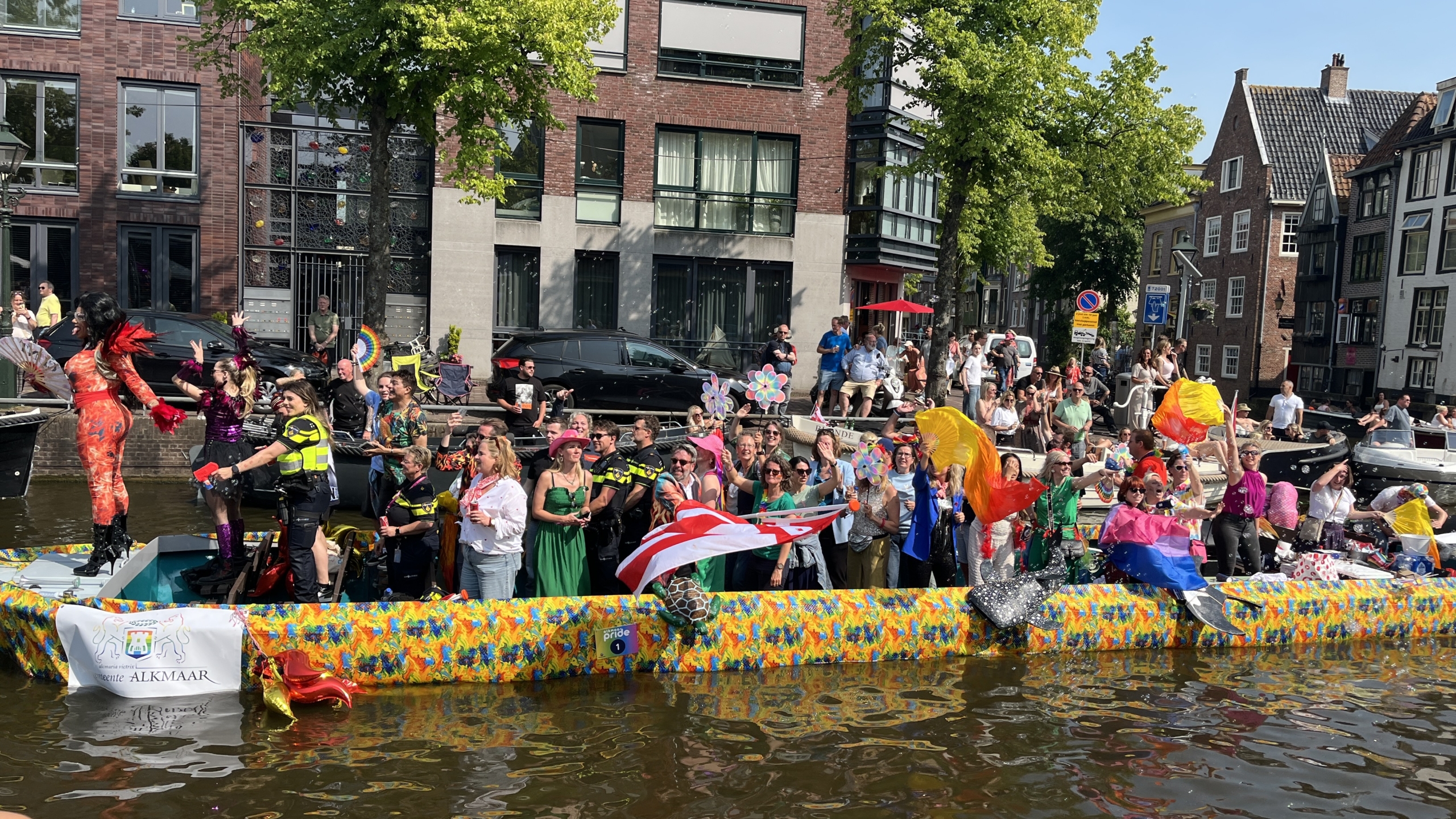 Een kleurrijke boot vol feestende mensen vaart door een kanaal, omringd door toeschouwers aan de oevers, tijdens een pride-parade in een stedelijke omgeving.