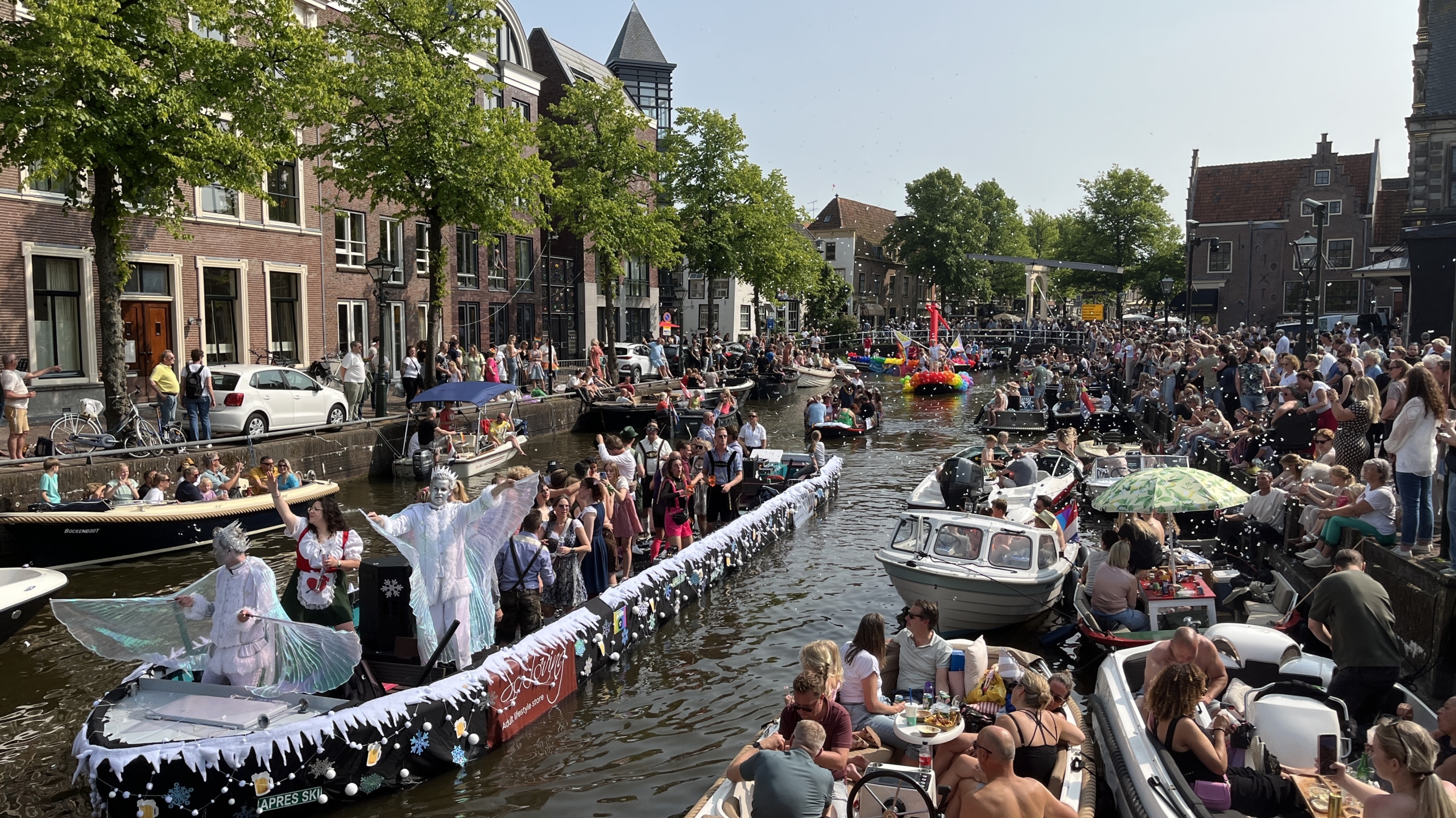 Een druk bezochte botenparade op een zonnige dag met mensen op en langs de gracht, waaronder een versierde boot met kostuumdragers.