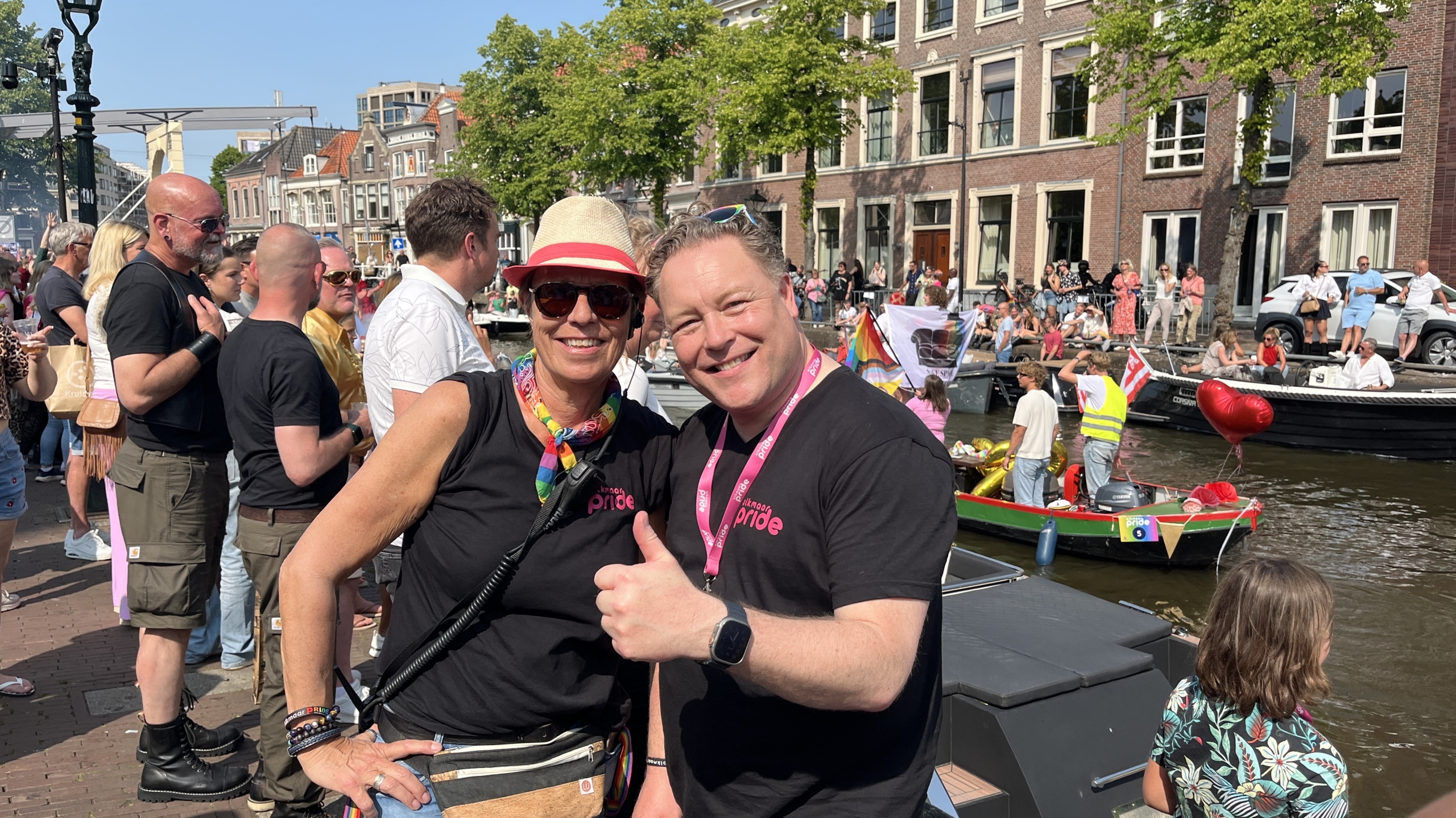 Twee mensen in Pride T-shirts poseren voor de camera met een duim omhoog, omgeven door een menigte langs een gracht met boten, tijdens een zonnige dag. Een van de boten heeft regenboogvlaggen, en er is een rode ballon in de vorm van een hart te zien. Op de achtergrond zijn oude gebouwen zichtbaar.