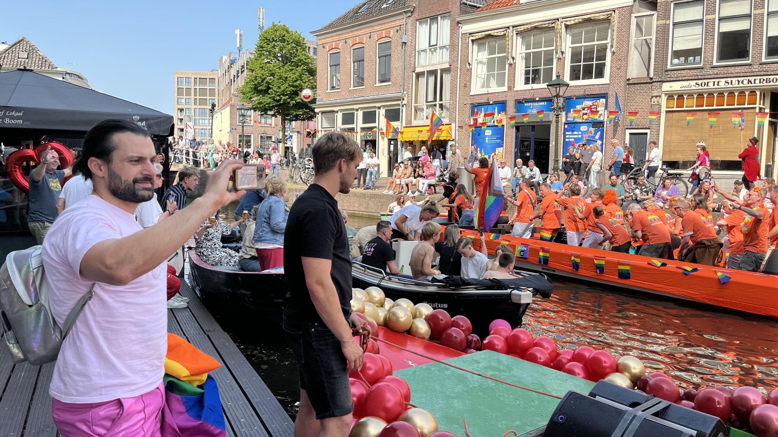 Een groep mensen in oranje T-shirts vaart op een versierde boot tijdens een kanalfestival, terwijl het publiek op de kade toekijkt en foto's maakt. Er hangen regenboogvlaggen en ballonnen in de omgeving.