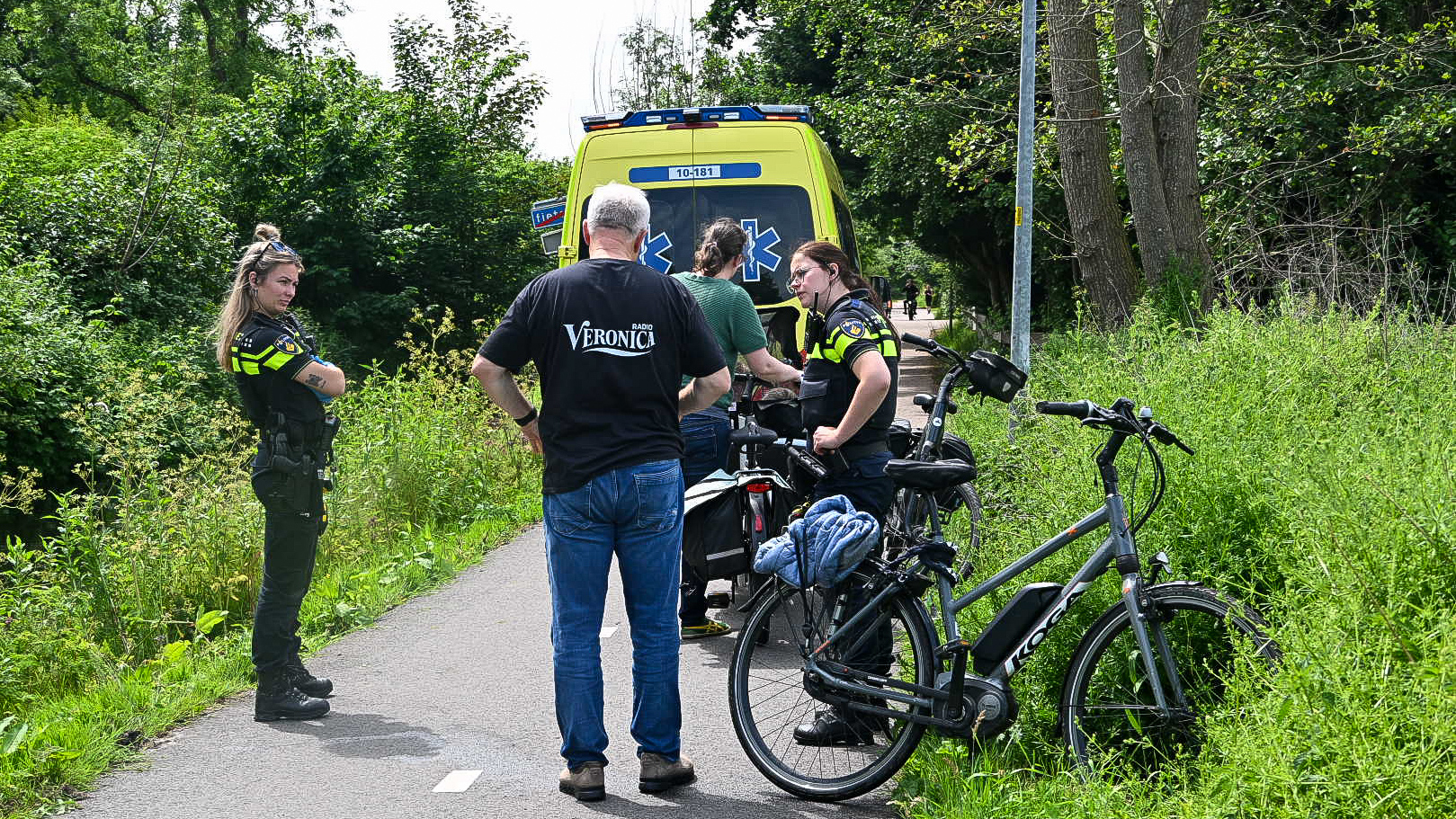 Twee politieagenten en een man staan naast een fietspad met een ambulance op de achtergrond, omringd door groen gebladerte.