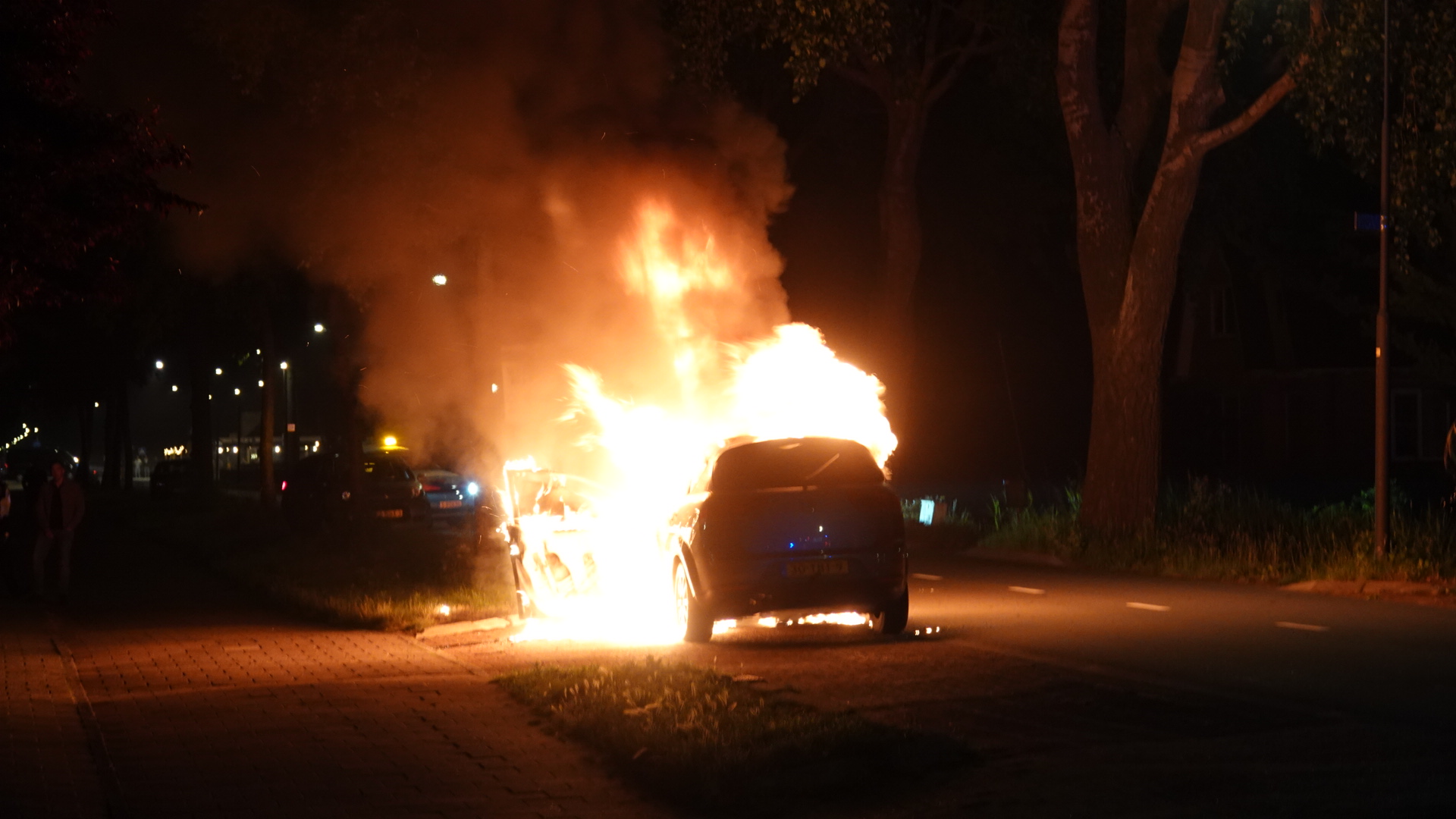 Auto die in brand staat langs een weg in de nacht, met grote vlammen en rookpluimen die opstijgen.