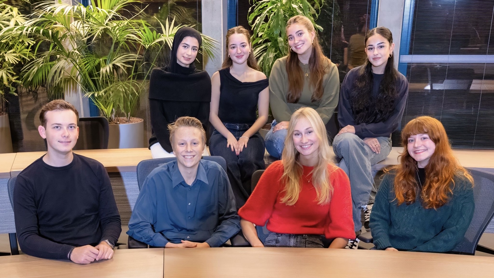 Een groep van acht jonge mensen posing in een kantooromgeving, met grote groene planten op de achtergrond.