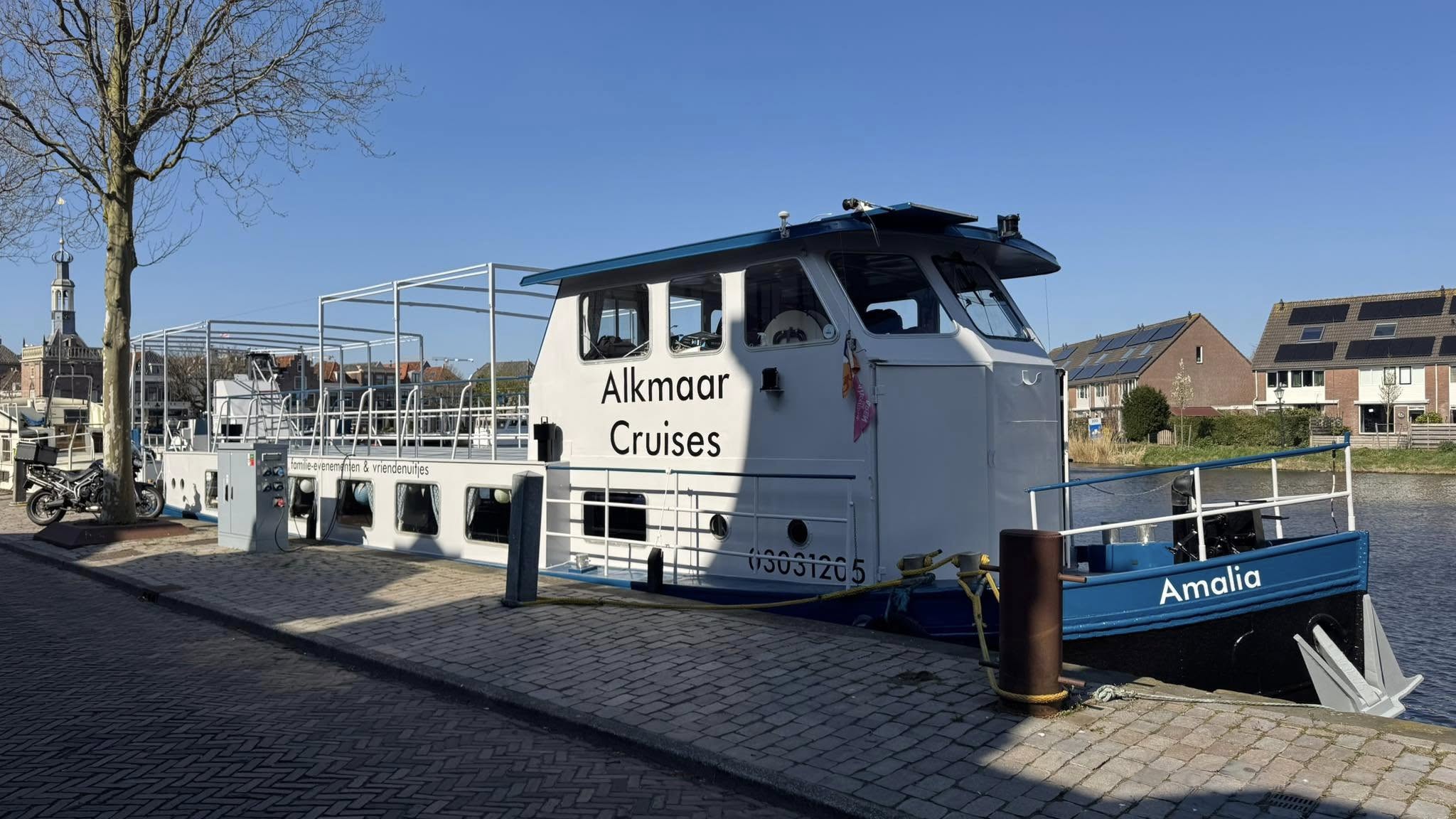 Een passagiersboot met de naam 'Amalia' van Alkmaar Cruises ligt aangemeerd aan een kade, met op de achtergrond een toren en woonhuizen met zonnepanelen.