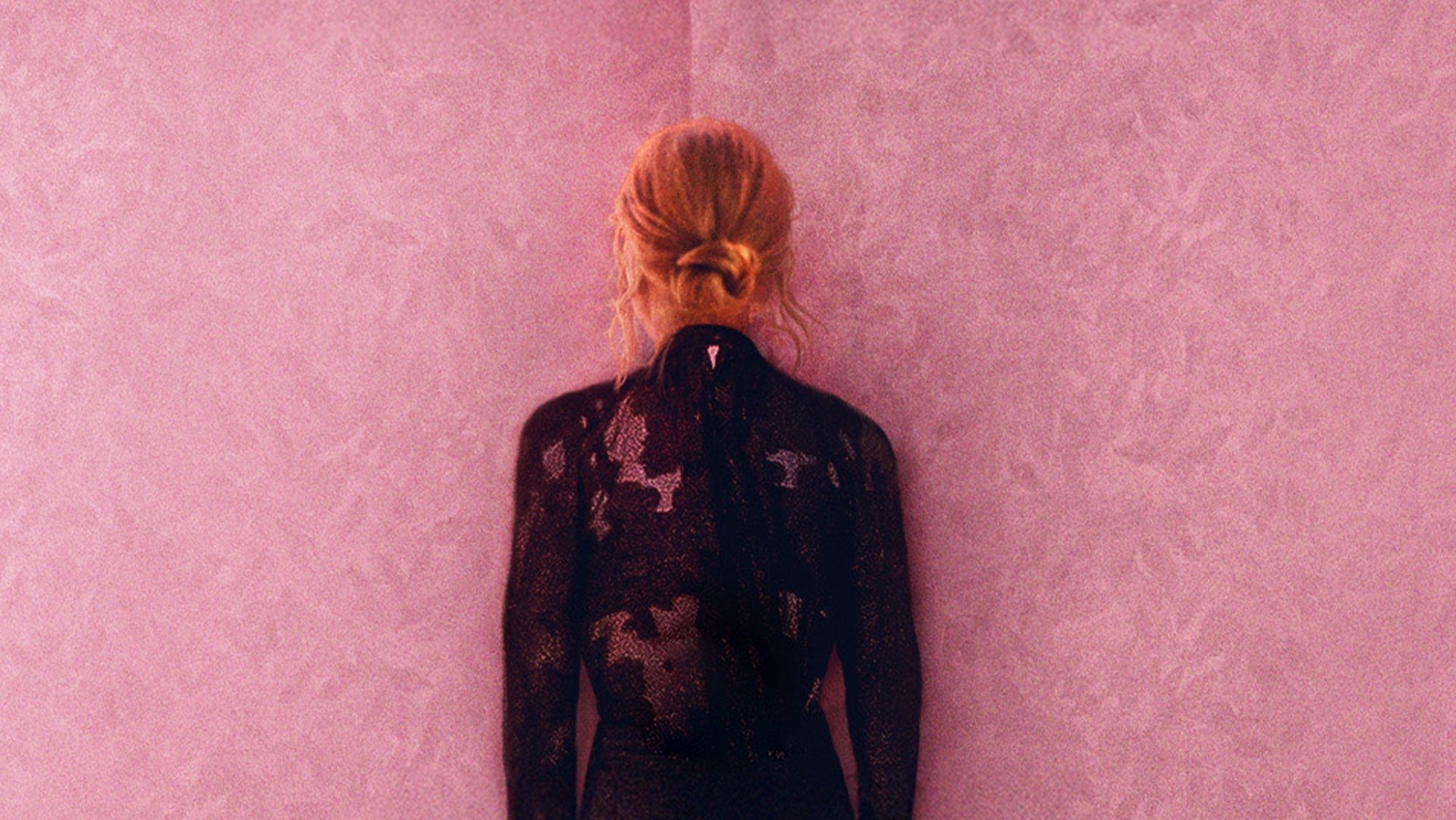 Een persoon met blond haar in een knot staat met de rug naar de camera tegen een roze muur, gekleed in een zwarte jurk met glitterpatroon.