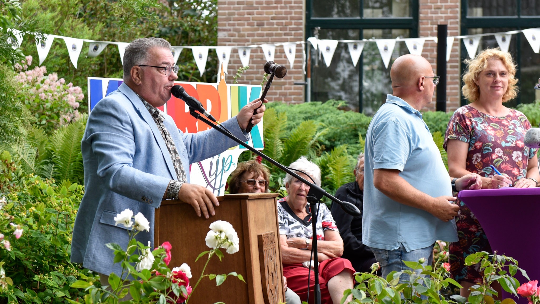 Een man met een blauwe blazer staat achter een spreekgestoelte, houdt een hamer vast en spreekt in een microfoon tijdens een evenement in een tuin. Op de achtergrond zijn mensen zittend op stoelen en een kleurrijk bord zichtbaar.
