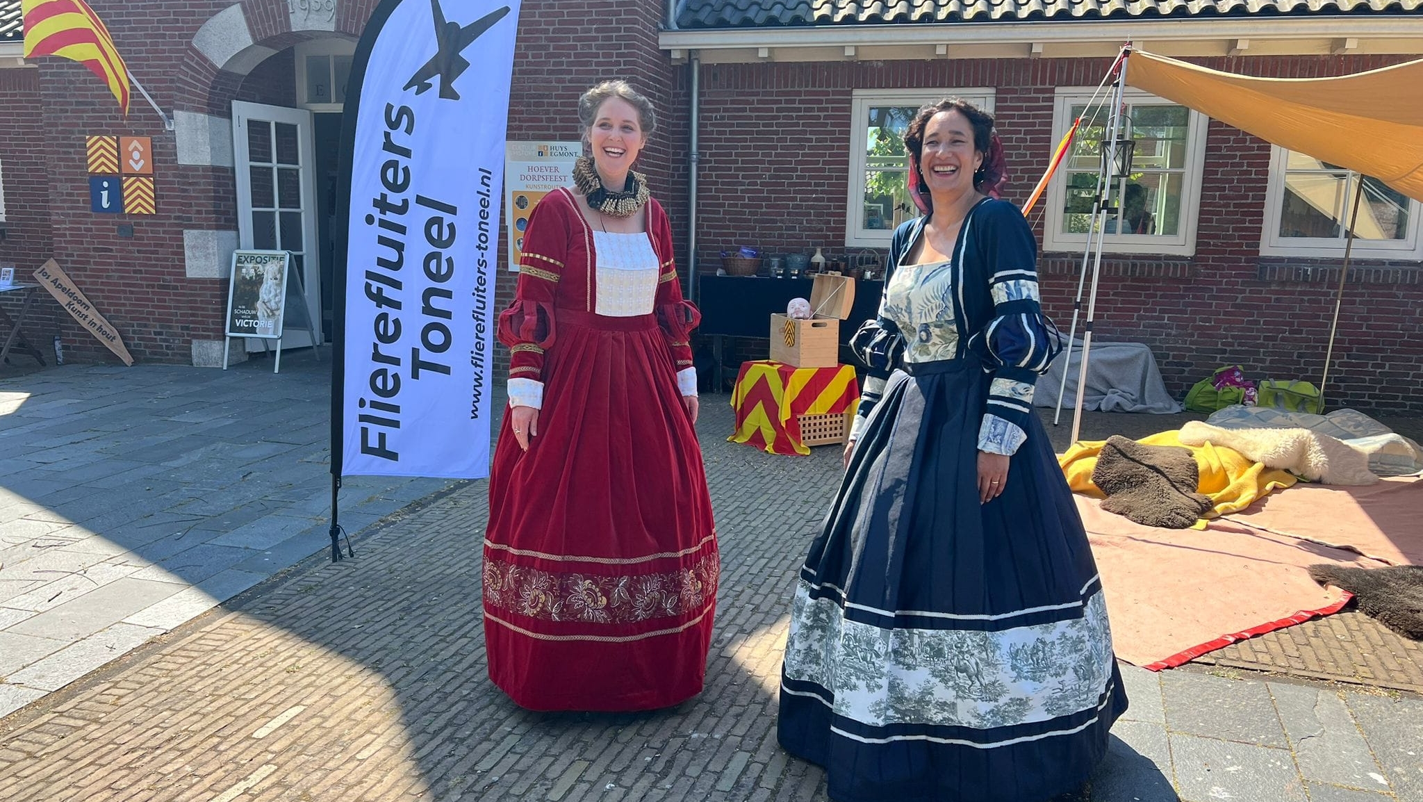 Twee vrouwen gekleed in historische jurken staan buiten naast een vlag met de tekst 'Flierefluiters Toneel'. Het gebouw op de achtergrond heeft bakstenen muren en decoratieve elementen.