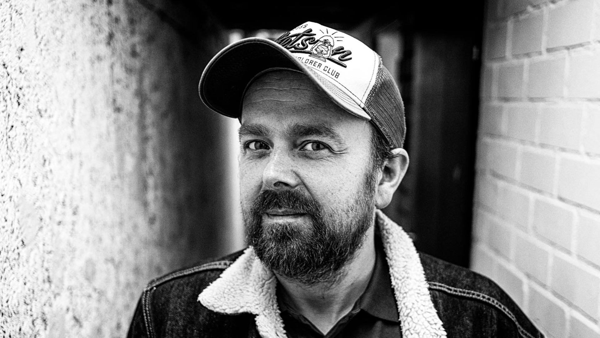 Zwart-wit portret van een man met een baard, een pet en een jas met een bontkraag. Hij staat tussen een stenen muur en een bakstenen muur, kijkend naar de camera.