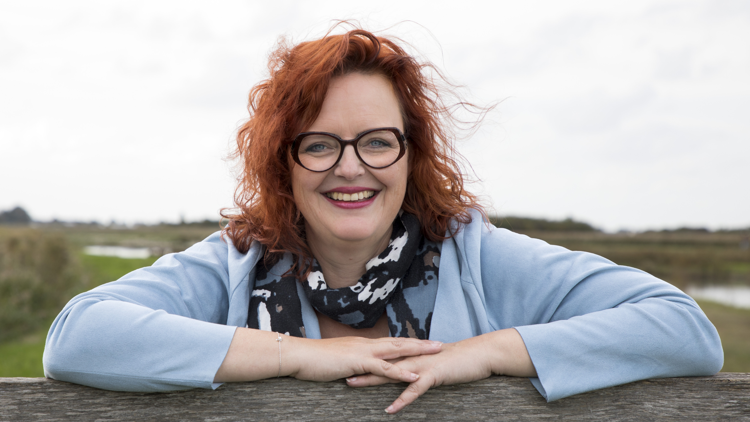 Vrouw met rood haar en een bril, leunend aan de rand van een houten hek, in een open veld met een rivier op de achtergrond.