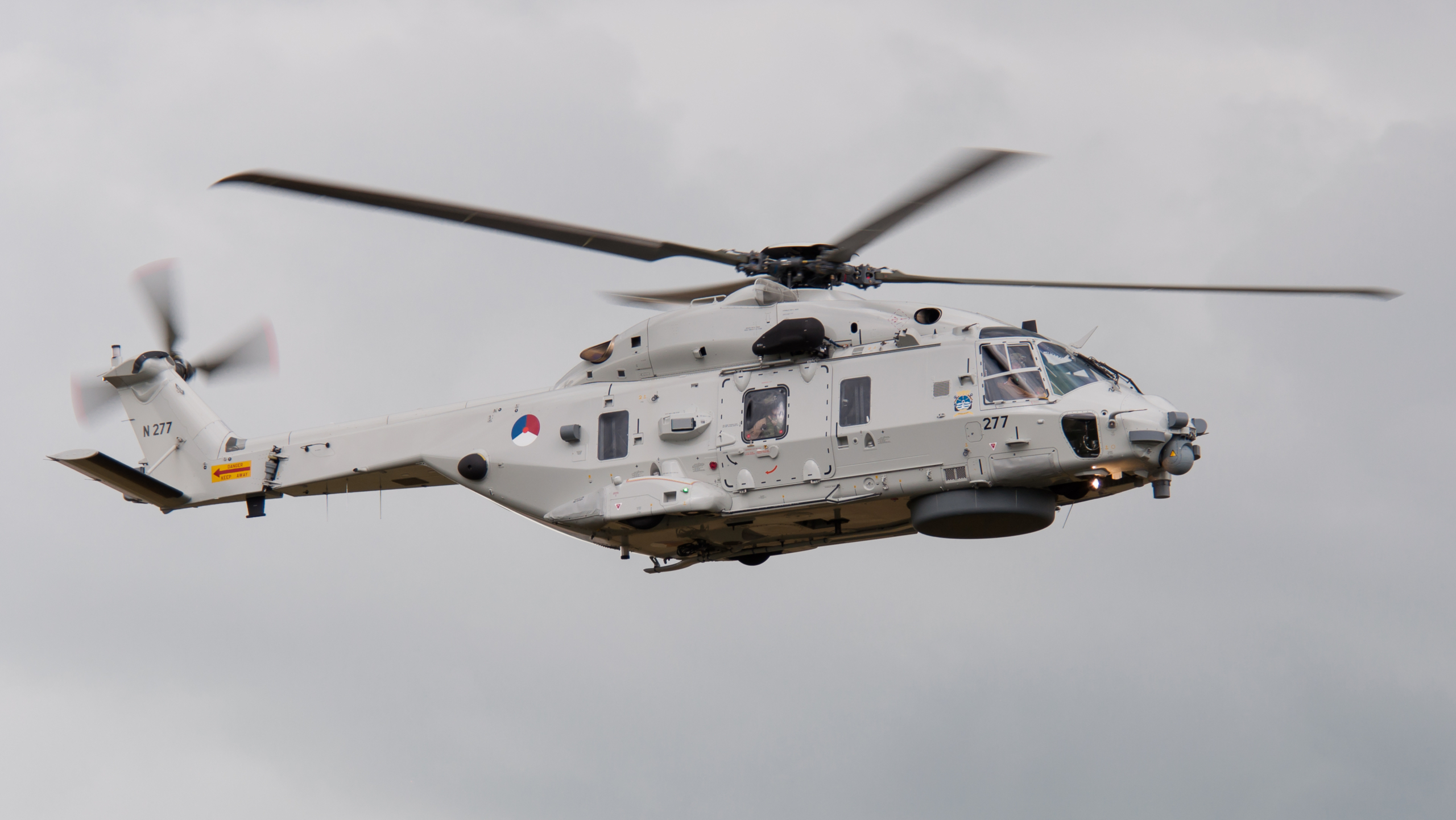Grijze militaire helikopter in de lucht met draaiende rotors, boven een bewolkte hemel.