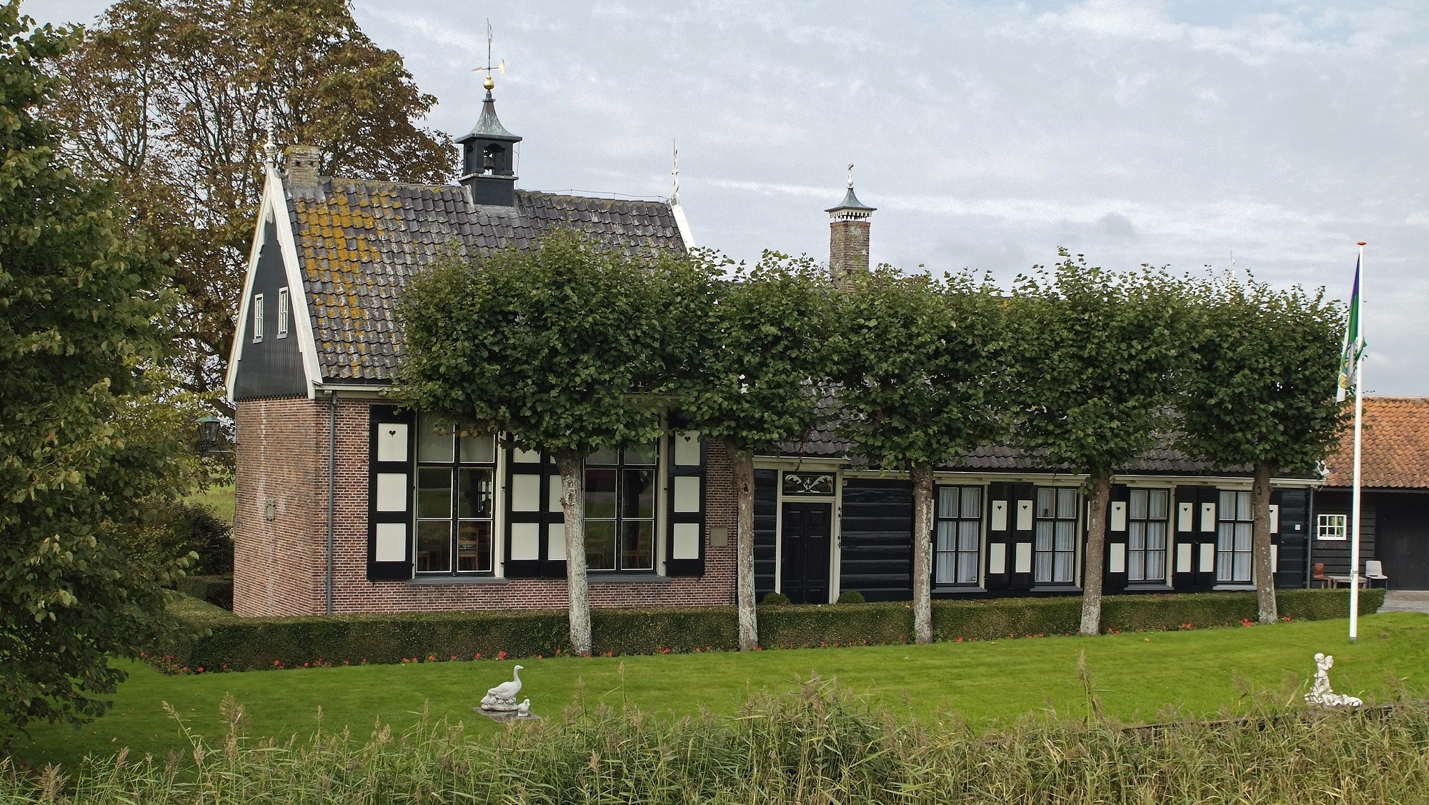 Een traditioneel Nederlands huis met stenen muren en een puntdak, omringd door een grasveld en bomen; op de voorgrond is een gracht zichtbaar.