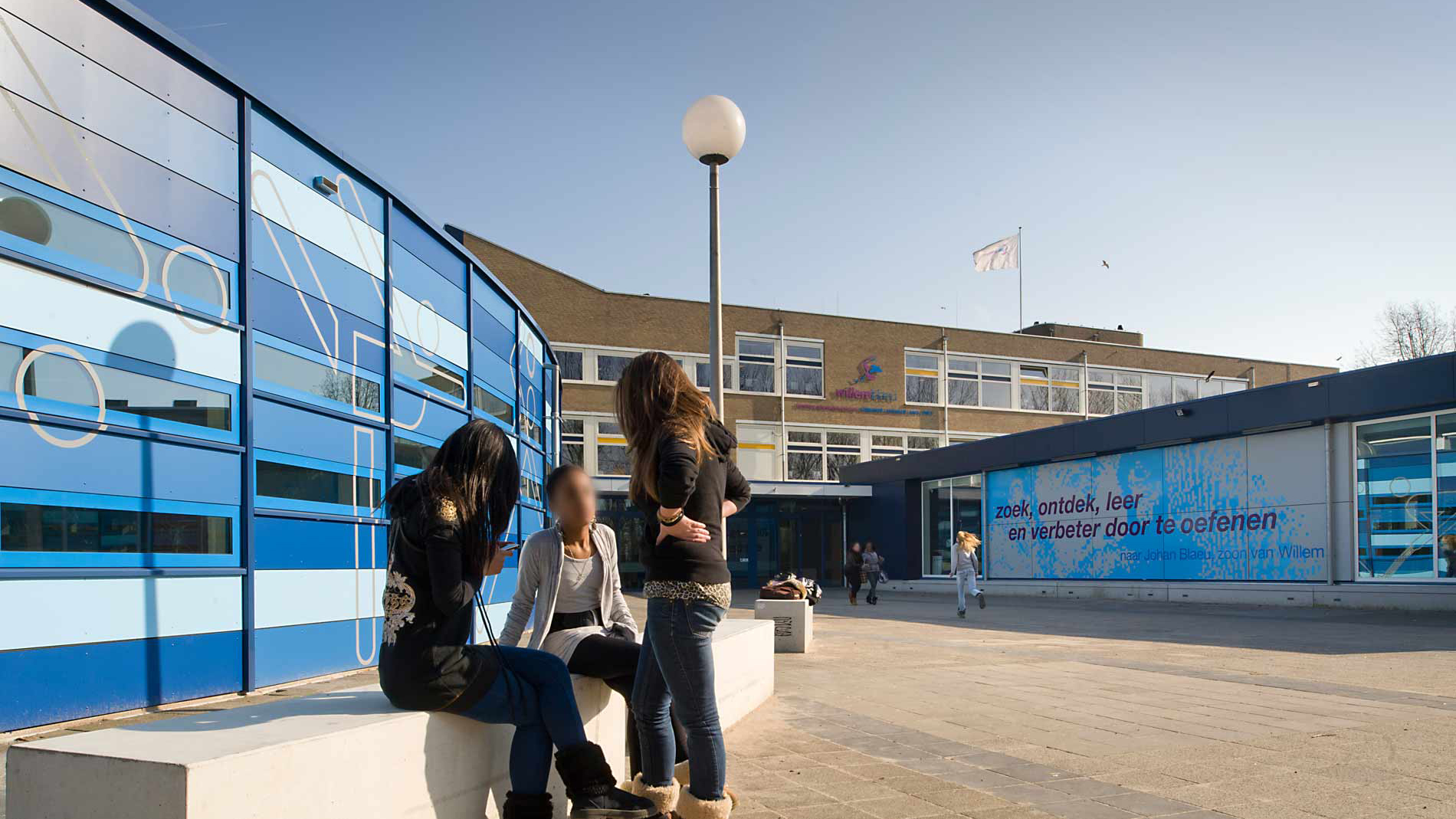 Drie mensen staan samen buiten voor een blauw gebouw met een glazen gevel. Het gebouw heeft een grote tekst op de muur met de boodschap "zoek, ontdek, leer en verbeter door te oefenen."