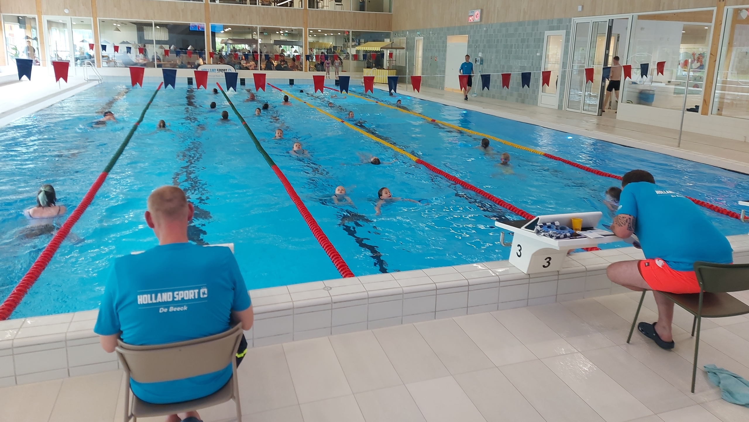 Binnenzwembad met meerdere zwemmers in verschillende banen. Twee personen in blauwe shirts zitten aan de rand van het zwembad met notities of apparatuur. Het zwembad heeft een houten plafond en glazen wanden die een tweede verdieping met uitzicht op het zwembad tonen.