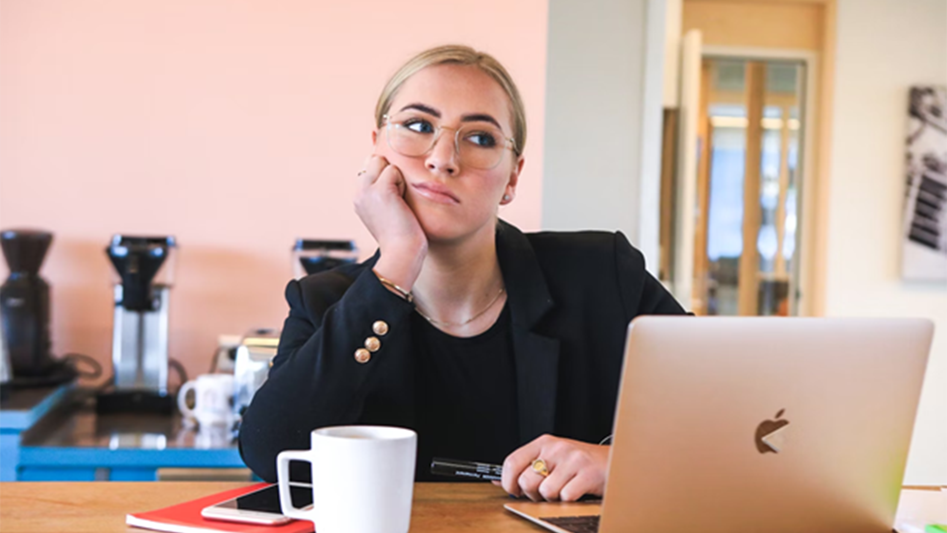 Vrouw met een bril en blond haar, geleund op haar hand, kijkt verveeld terwijl ze achter een laptop zit, met een mok en een notitieboekje op tafel.