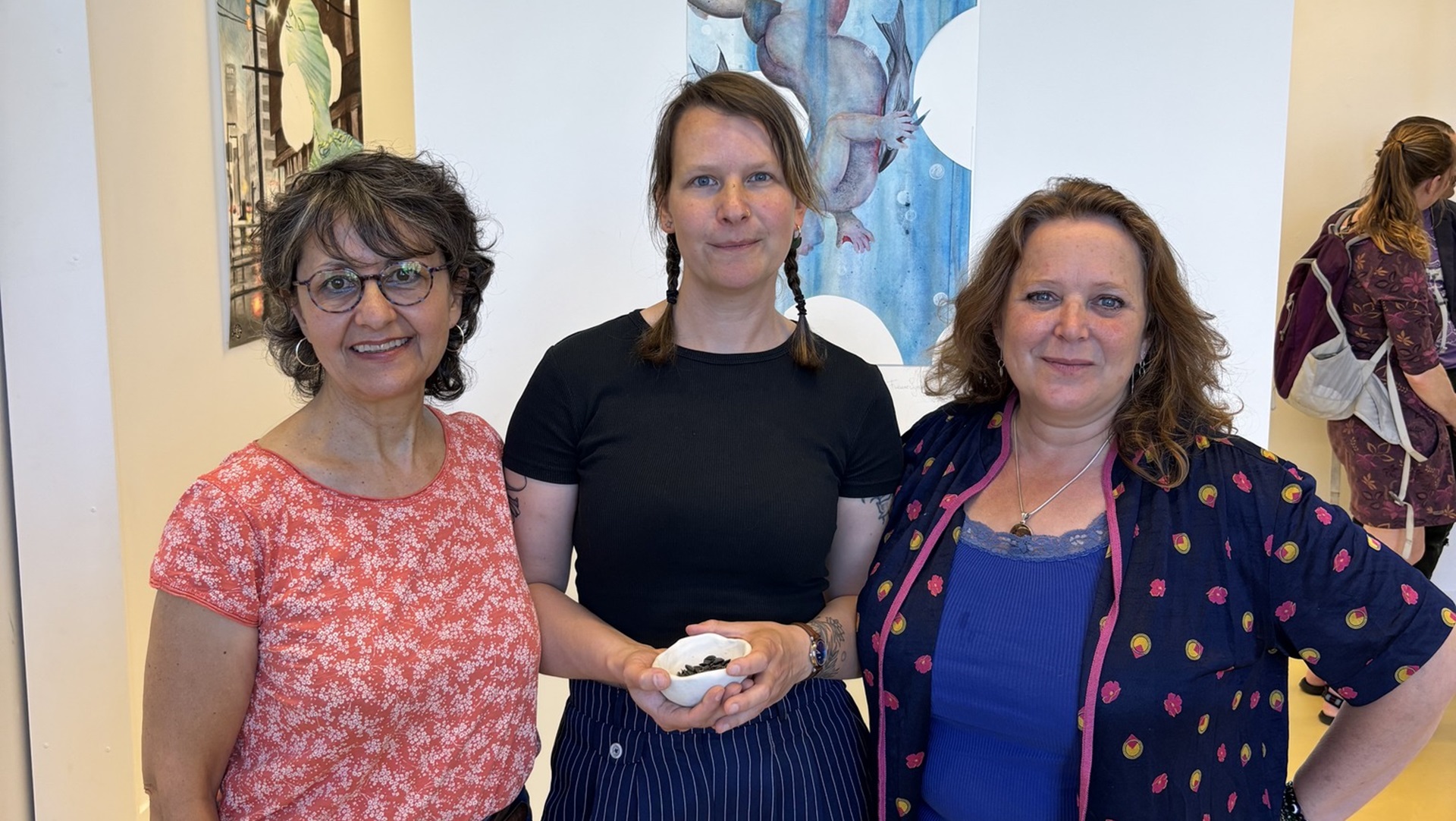 Drie vrouwen poseren samen binnen in een gebouw met kunstwerken op de achtergrond; de middelste vrouw houdt een klein wit schoteltje vast.