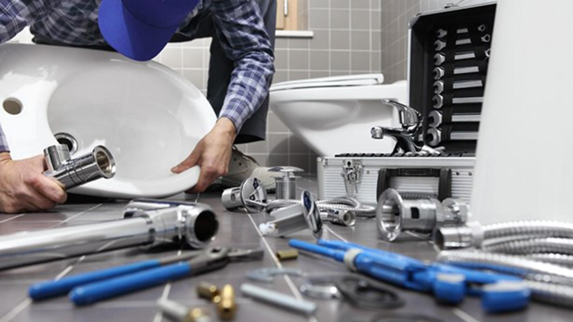 Een loodgieter werkt in een badkamer en bevestigt een witte wastafel; gereedschap en onderdelen liggen verspreid op de tegelvloer.