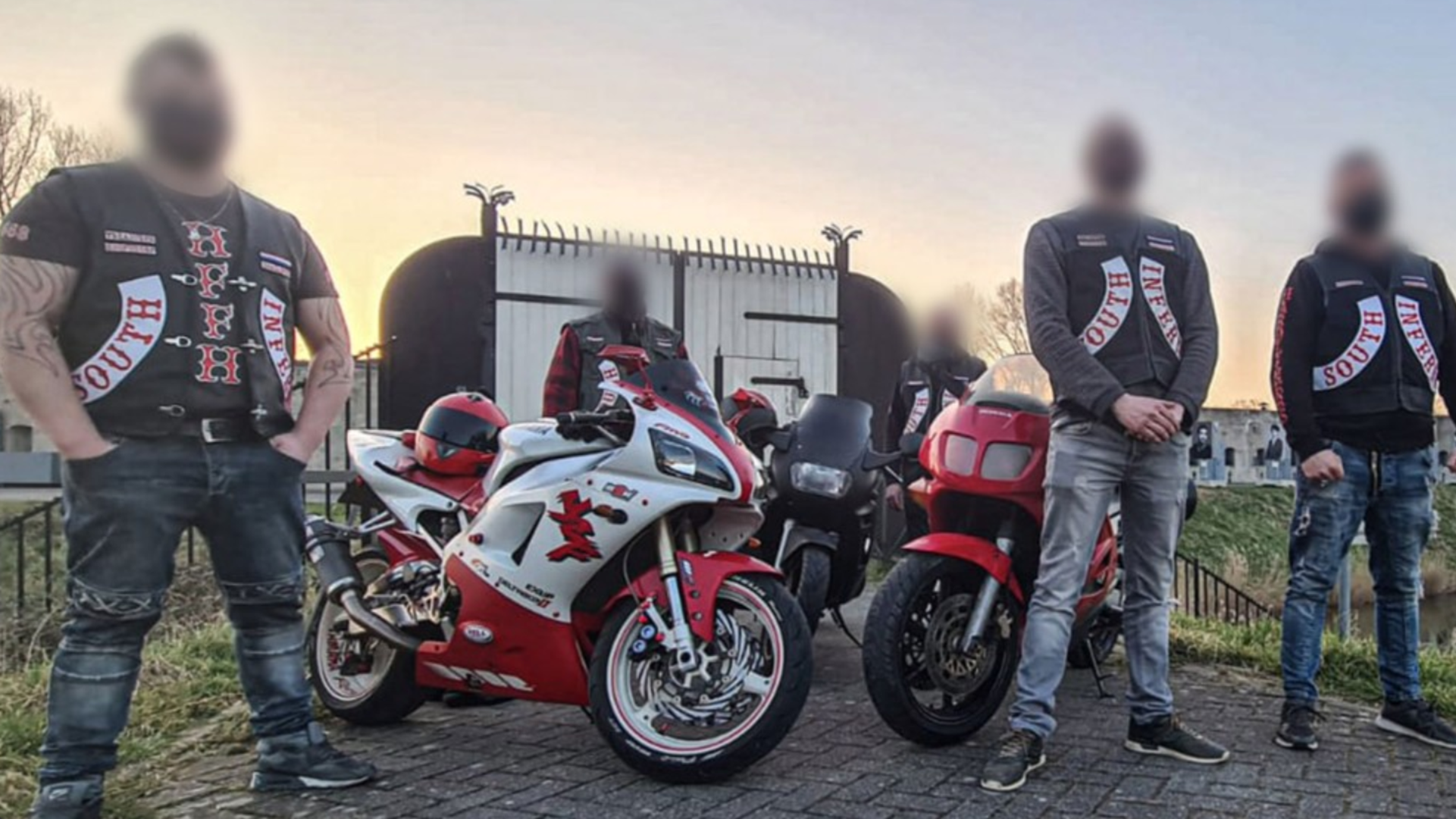 Vijf mannen met ingevagen gezichten staan bij drie motorfietsen, terwijl ze jassen dragen met de tekst 'South Inf' op de schouders, voor een ijzeren hek.