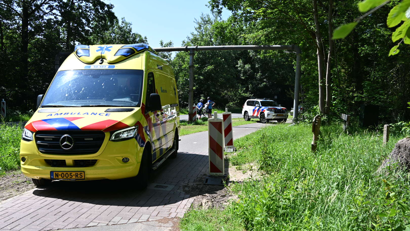 Een gele ambulance en een politieauto staan op een weg bij een groen bosrijk gebied, omgeven door gras en bomen.