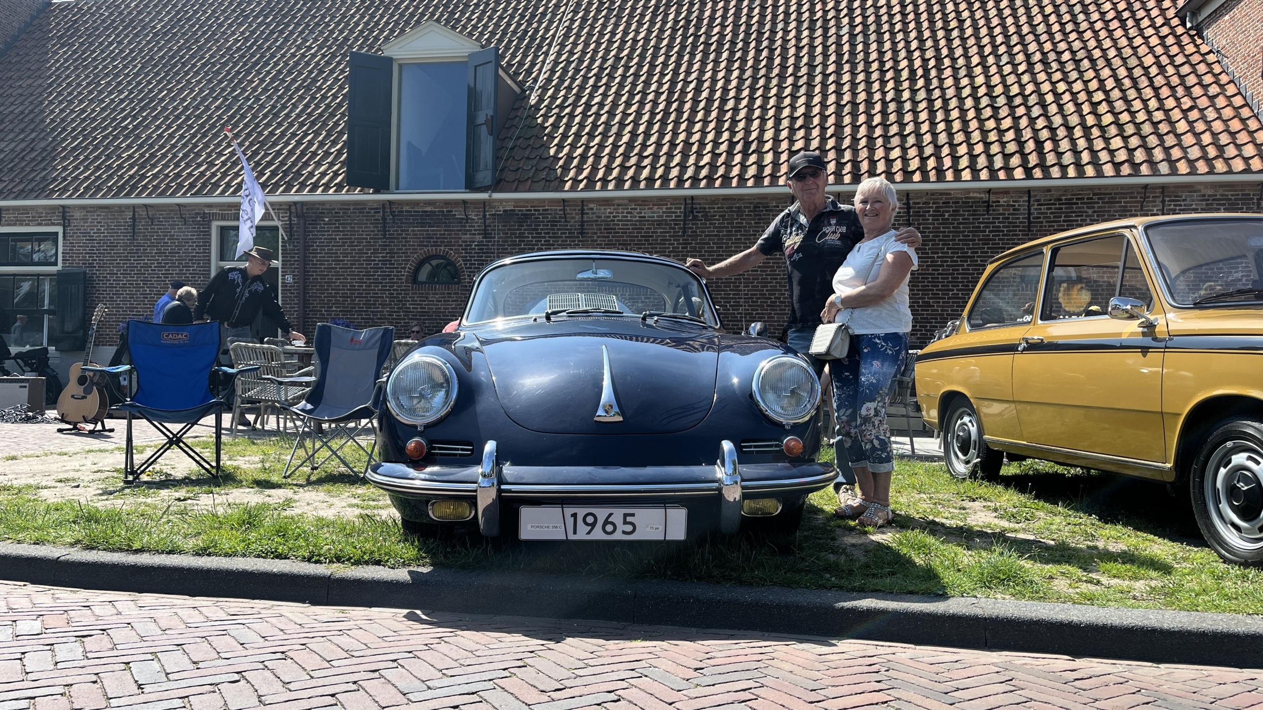 Twee mensen poseren naast een blauwe klassieke auto uit 1965, geparkeerd naast een geel voertuig, voor een bakstenen gebouw met een hellend pannendak.