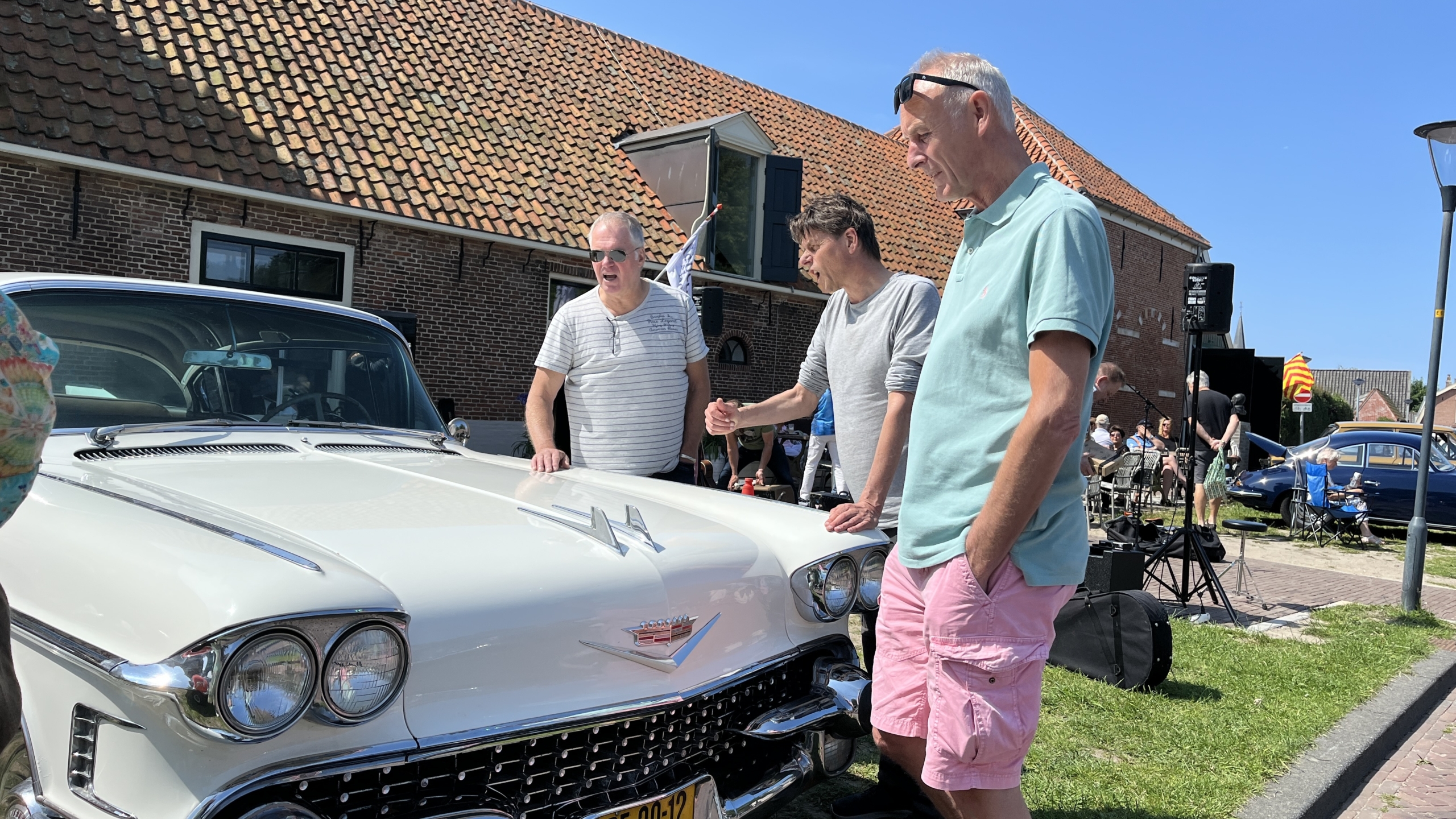 Drie mannen bekijken een witte klassieke auto met twee koplampen en een Cadillac-embleem op de motorkap, tegen de achtergrond van een bakstenen gebouw met een rood pannendak. Andere auto's en mensen zijn zichtbaar in de omgeving op een zonnige dag.