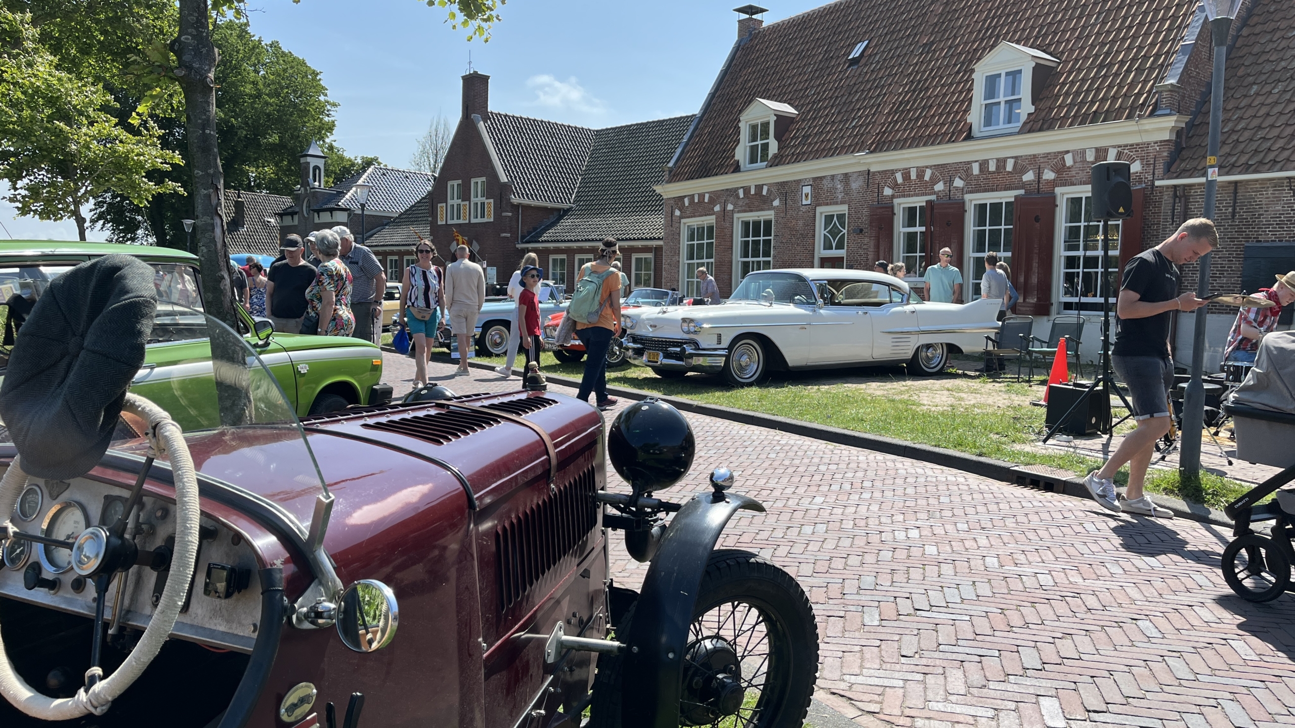 Oldtimers en klassieke auto's tentoongesteld op een zonnig plein met mensen die eromheen lopen, geflankeerd door huizen met roodbruine daken in een dorpachtige setting.