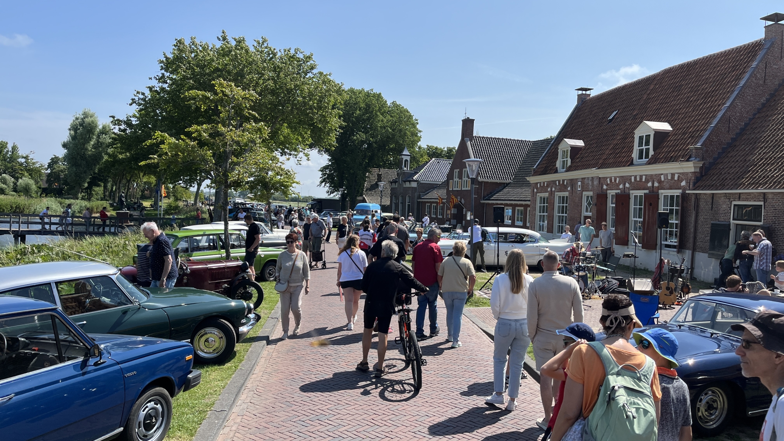 Mensen lopen langs een rij klassieke auto's die geparkeerd staan langs een geplaveide straat naast traditionele huizen en een groen parklandschap.