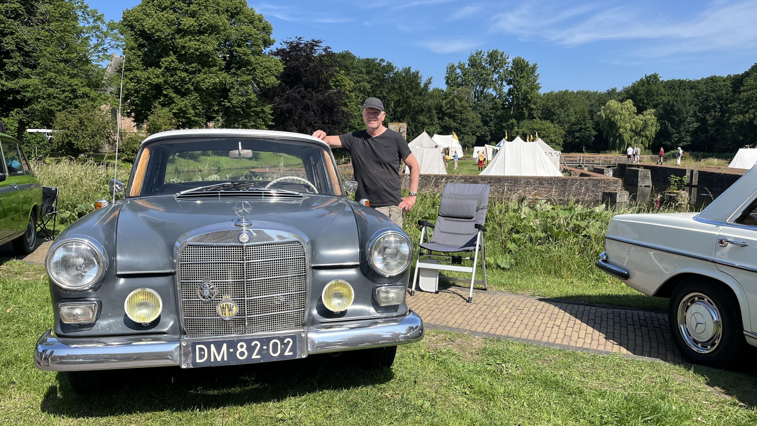 Man leans on a classic Mercedes-Benz car parked on a grassy area, with tents and trees in the background under a clear blue sky.