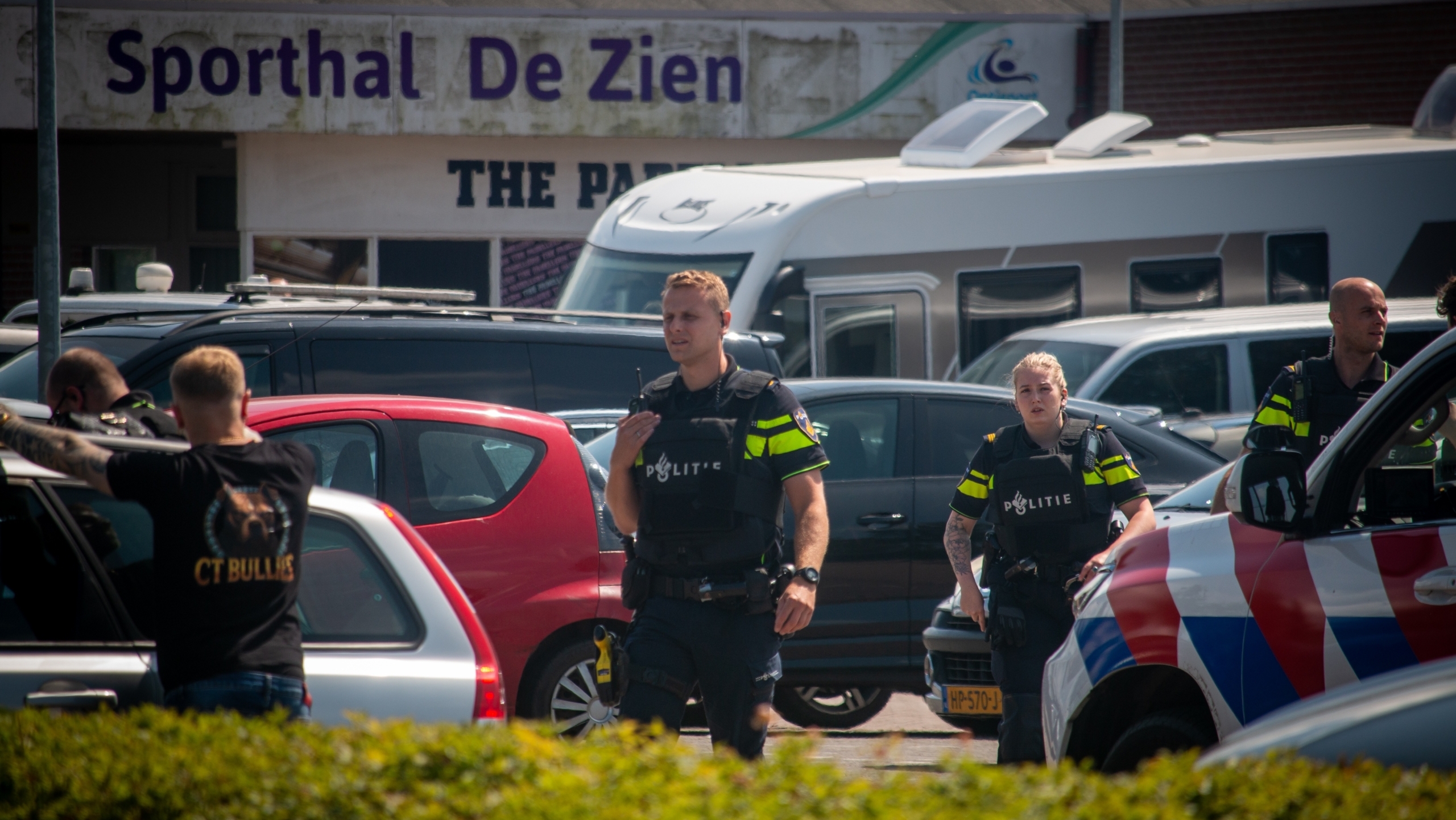 Politieagenten in uniform staan op een parkeerplaats voor een sporthal genaamd "Sporthal De Zien". Er zijn meerdere voertuigen, waaronder een politieauto met rood-wit-blauwe strepen en een camper, zichtbaar op de achtergrond.