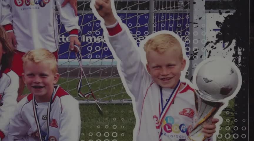 Twee jonge jongens in voetbaluniformen, de jongen rechts houdt een trofee omhoog en glimlacht trots; er is een voetbaldoel op de achtergrond.
