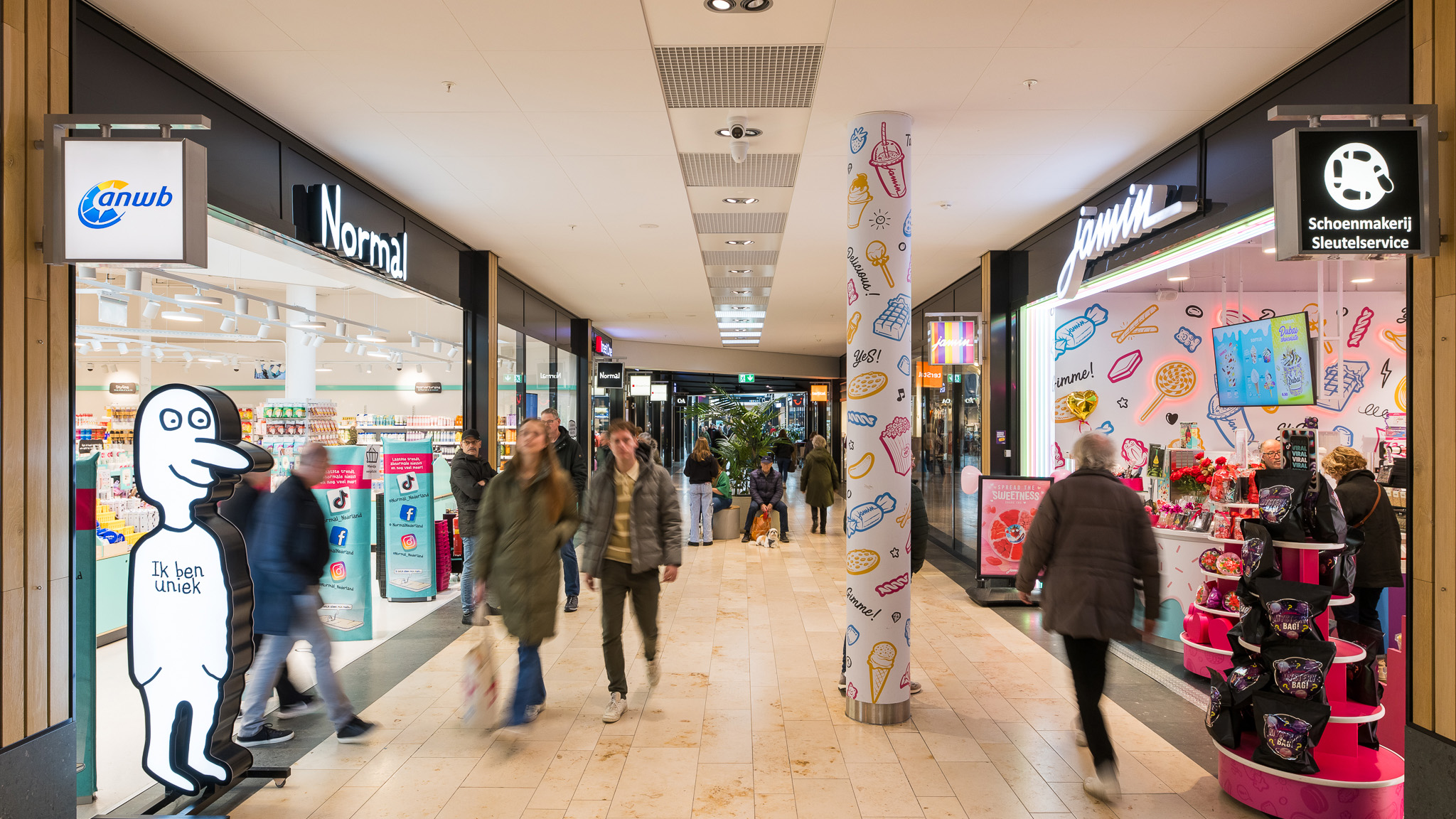 Mensen lopen door een winkelcentrum met winkels zoals Normal en Jamin aan weerszijden. Een opvallende zuil is versierd met kleurrijke tekeningen van voedsel zoals ijsjes. Een bord bij Normal heeft een cartoonfiguur met de tekst 'Ik ben uniek'.