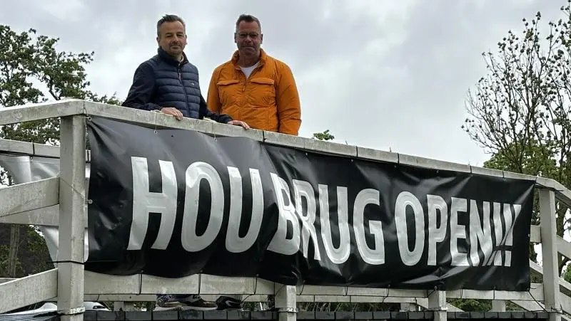 Twee mannen staan naast een houten reling met een zwart spandoek waarop 'HOU BRUG OPEN!!!' staat, onder een bewolkte hemel.