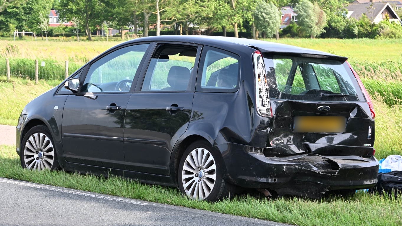 Een zwarte auto met aanzienlijke schade aan de achterkant staat geparkeerd op de berm naast een rustige weg, met gras en bomen op de achtergrond; enkele tassen liggen naast de auto.