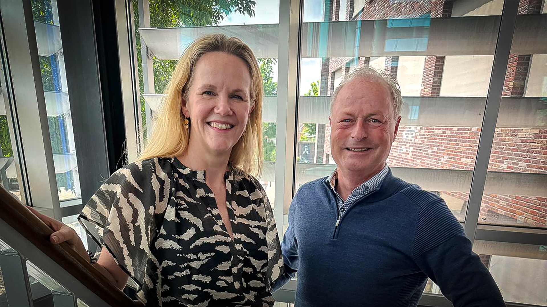 Een vrouw en een man staan glimlachend op een trap voor een glazen wand met zicht op bomen en bakstenen muren.